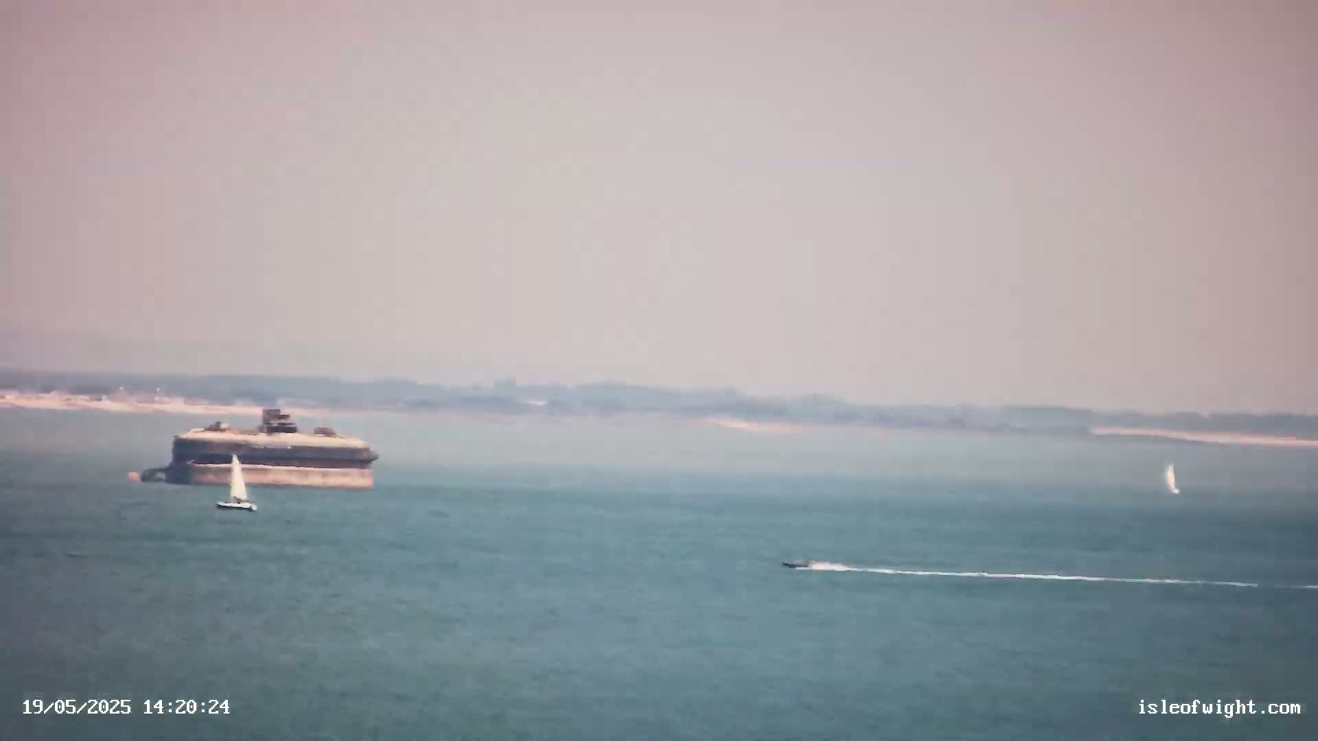 A hazy day shows a large, dark structure in the water with a sailboat nearby, and a small motorboat farther away, all against a background of land.
