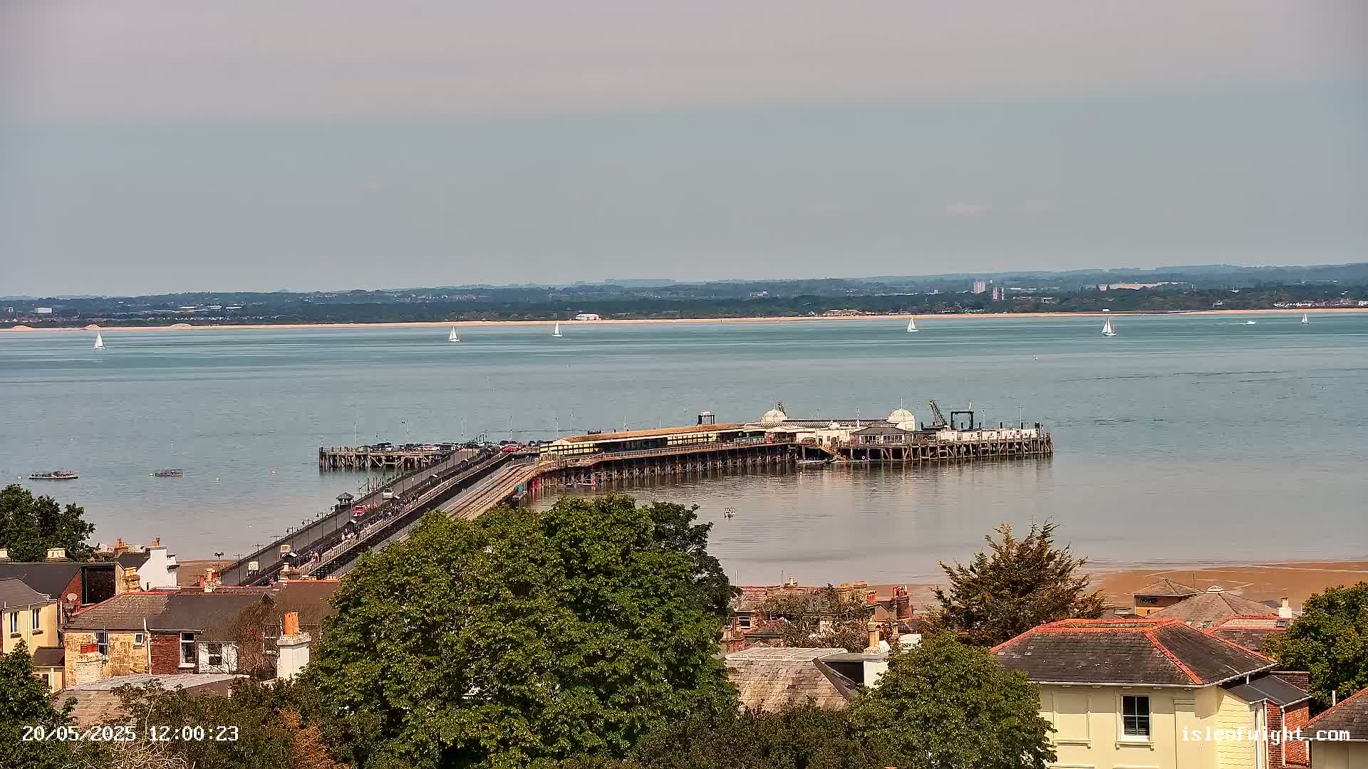 A long pier extends into a calm body of water under a mostly clear sky, with several sailboats visible in the distance and buildings and trees in the foreground.
