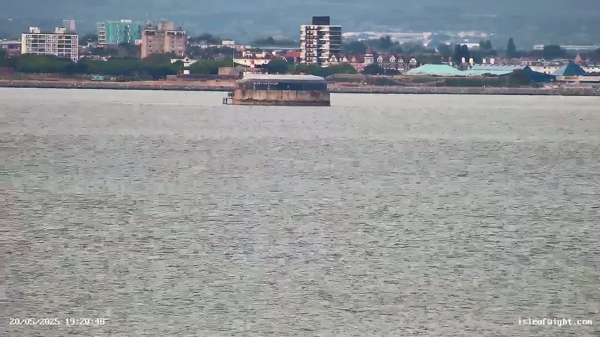 A low-lying, rectangular structure sits in a calm body of water in front of a city skyline under an overcast sky.