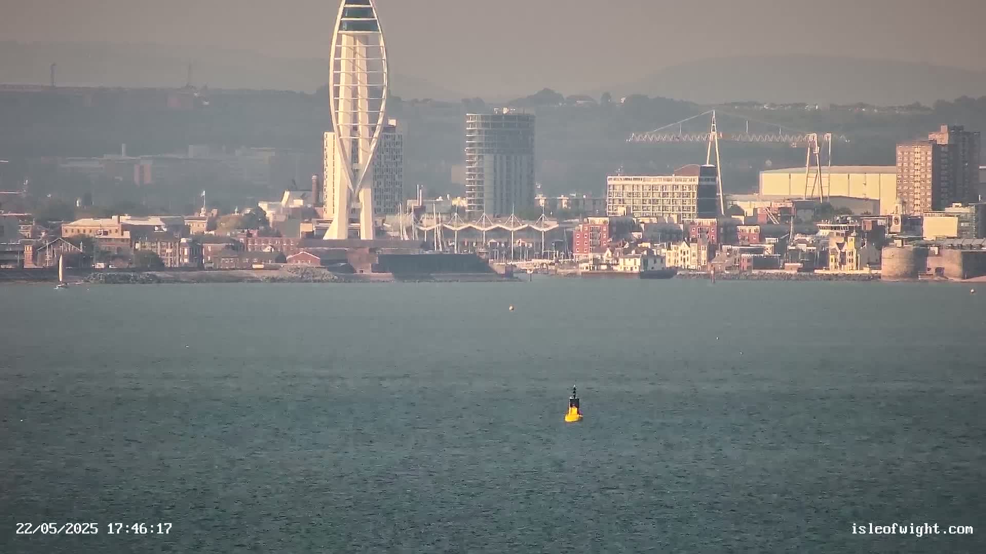A hazy view of a city skyline with a tall, slender tower, seen across a calm body of water with a single yellow buoy.