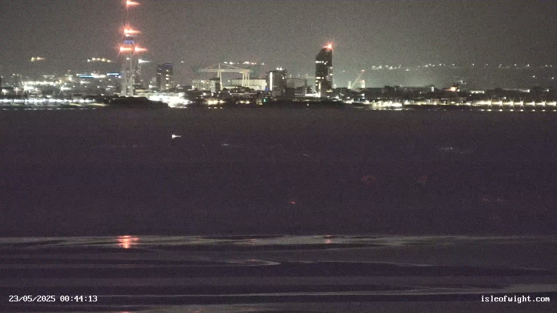 A nighttime view across dark water shows a city skyline with illuminated buildings and structures, under a dark sky.