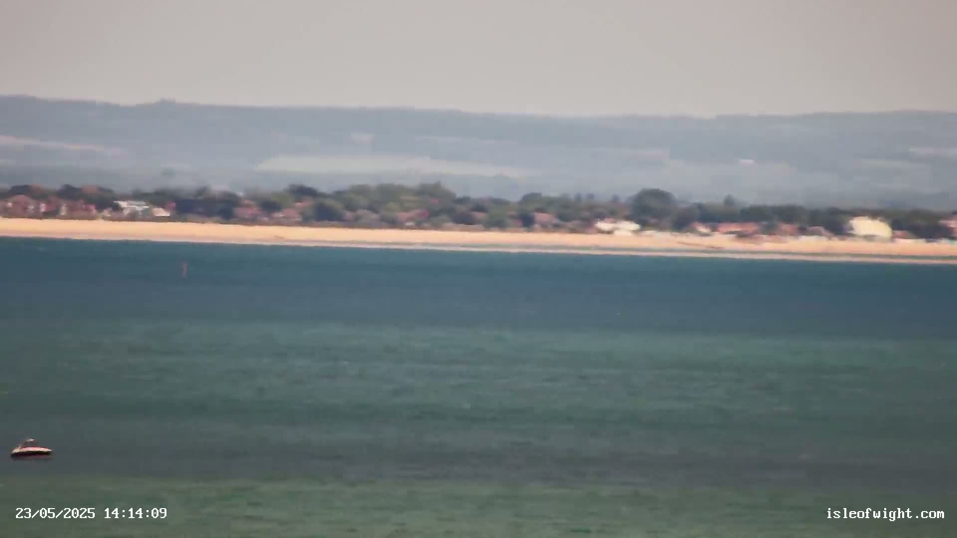 A small boat floats on a calm, teal sea in front of a sandy beach and distant hazy hills.