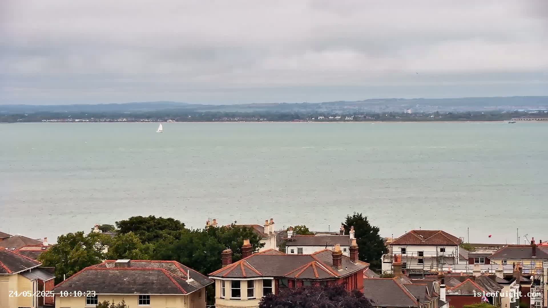 A sailboat is visible on a calm, gray body of water, overlooking a collection of houses with gray and red roofs under a cloudy sky.