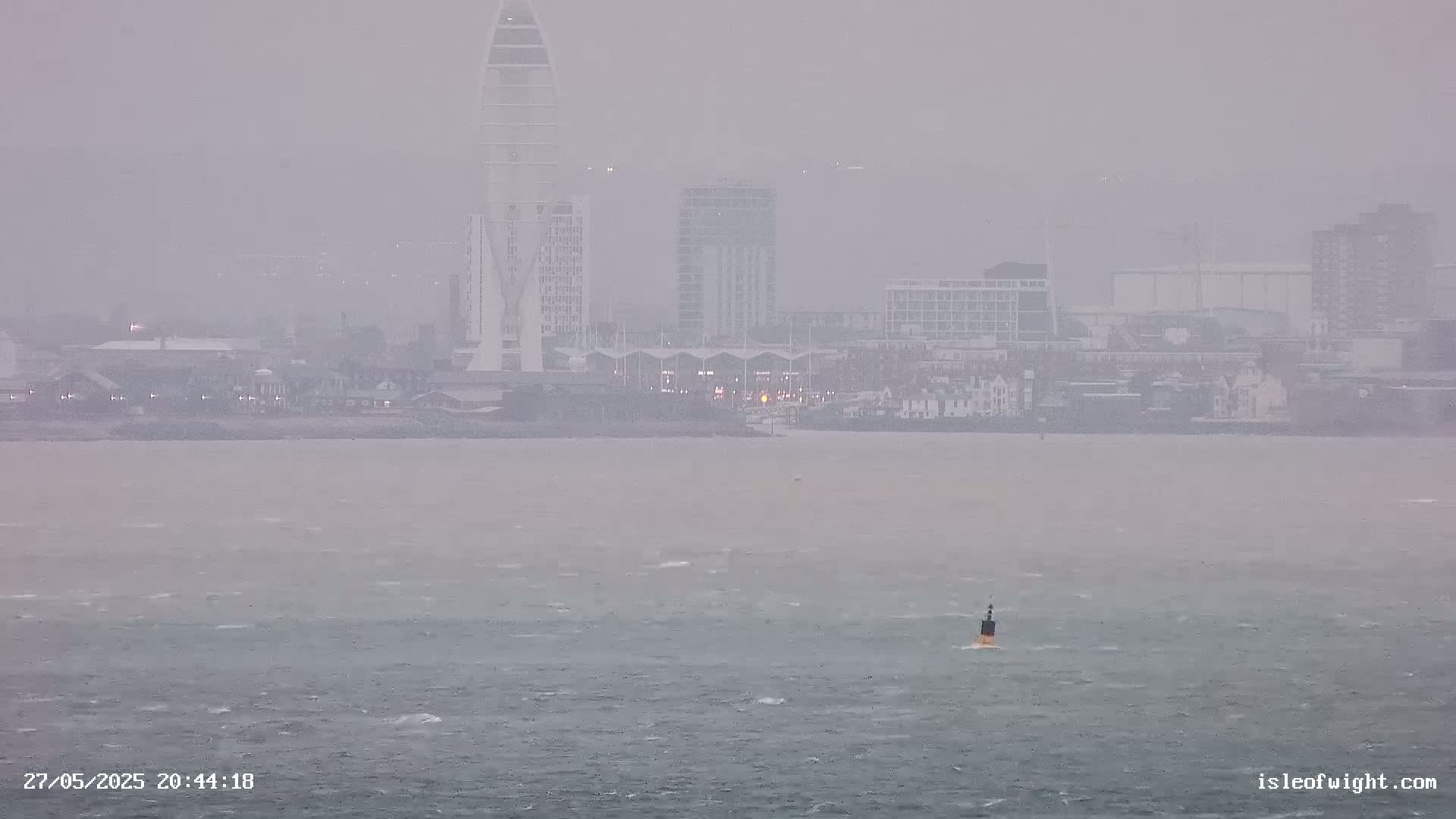 A distant cityscape with several buildings is partially obscured by heavy rain and wind over a choppy body of water with a single buoy visible.