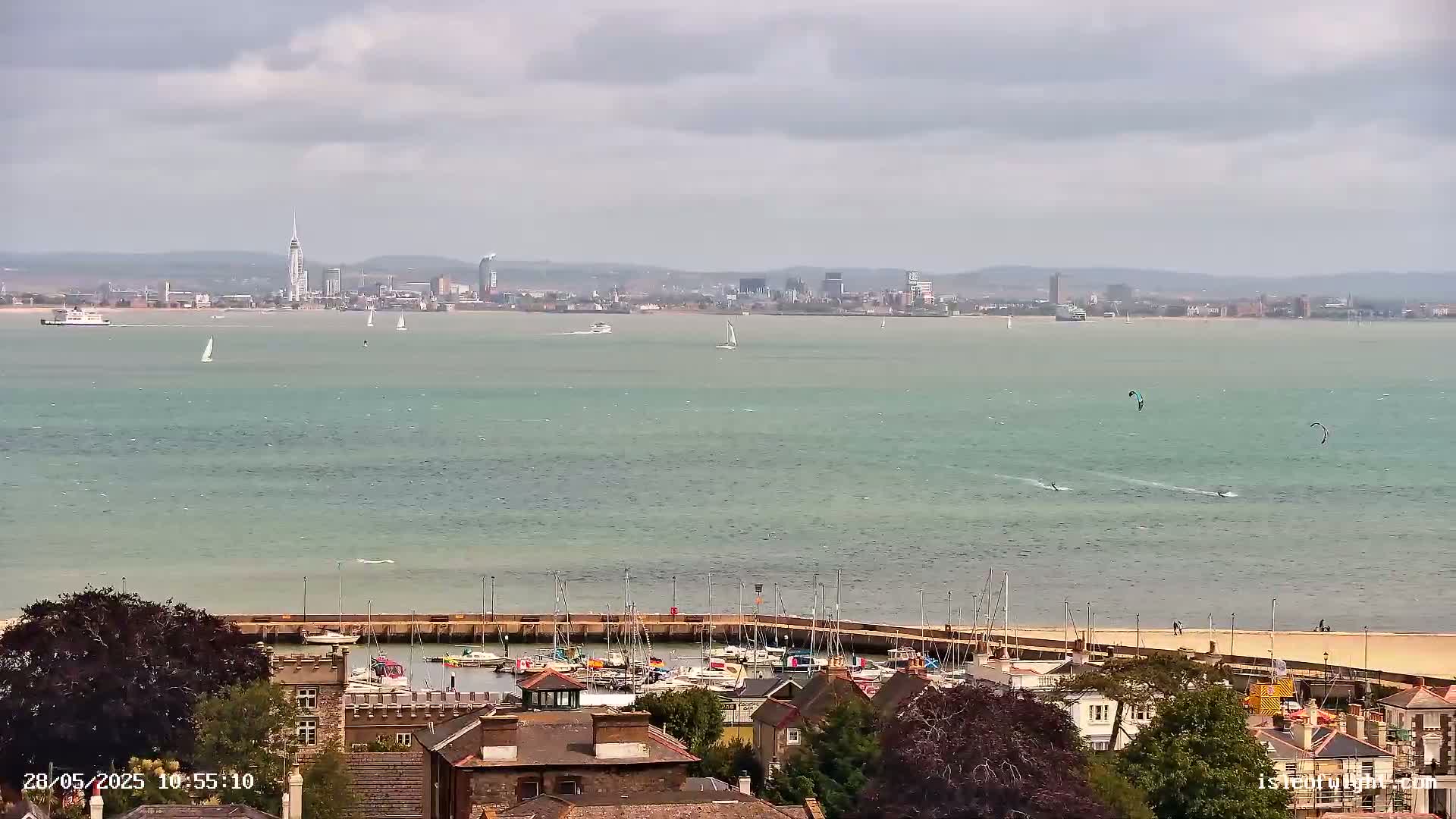 A seaside town with a marina is viewed from above on a partly cloudy day, overlooking a body of water with sailboats, kite surfers, and a distant city skyline.