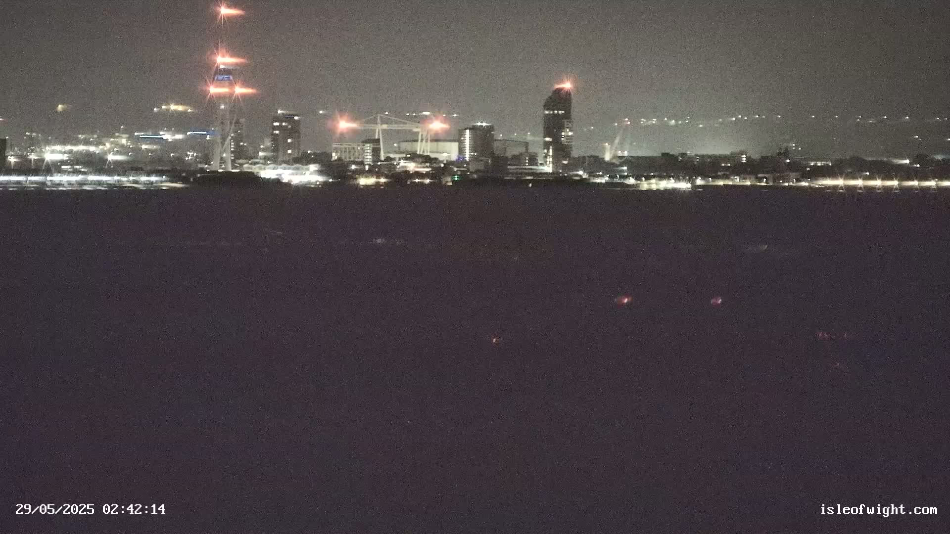 A nighttime view of a city skyline across a dark body of water, illuminated by various lights.