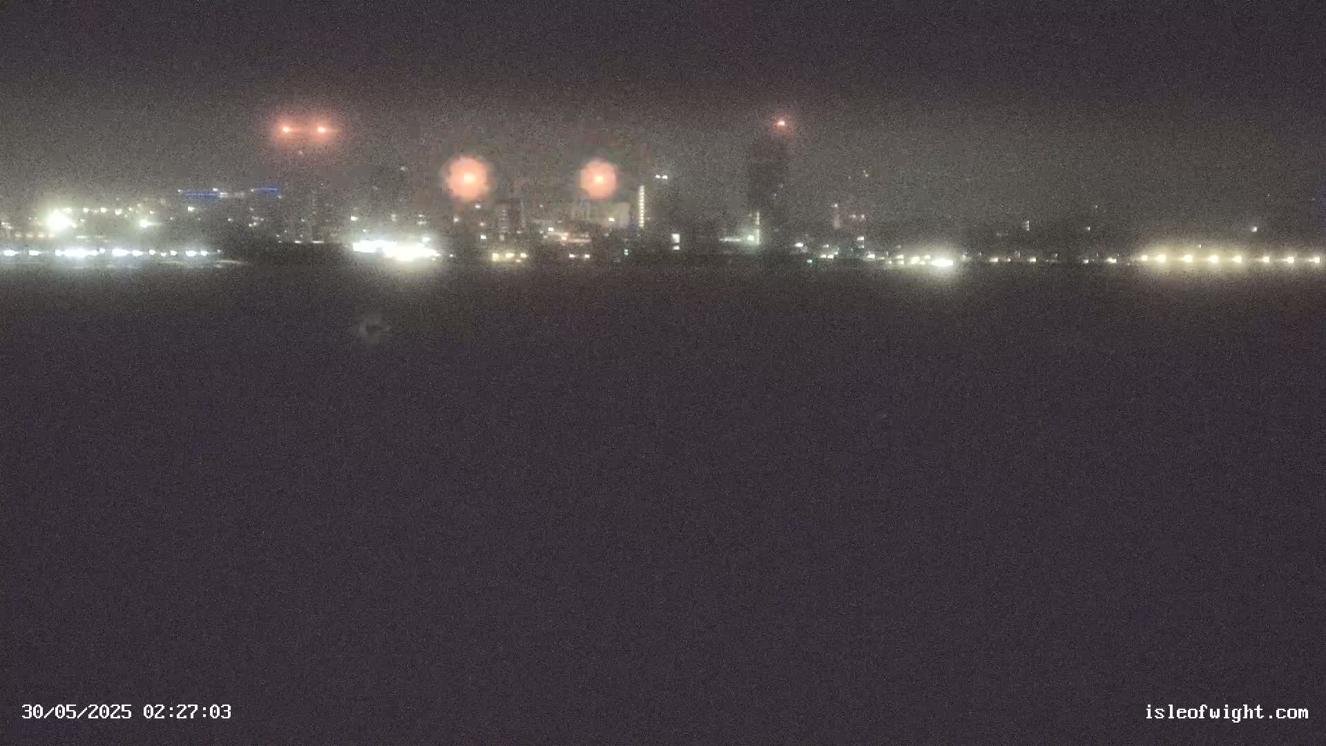 A hazy night scene shows a distant city skyline with lights illuminated, viewed from across a dark body of water.