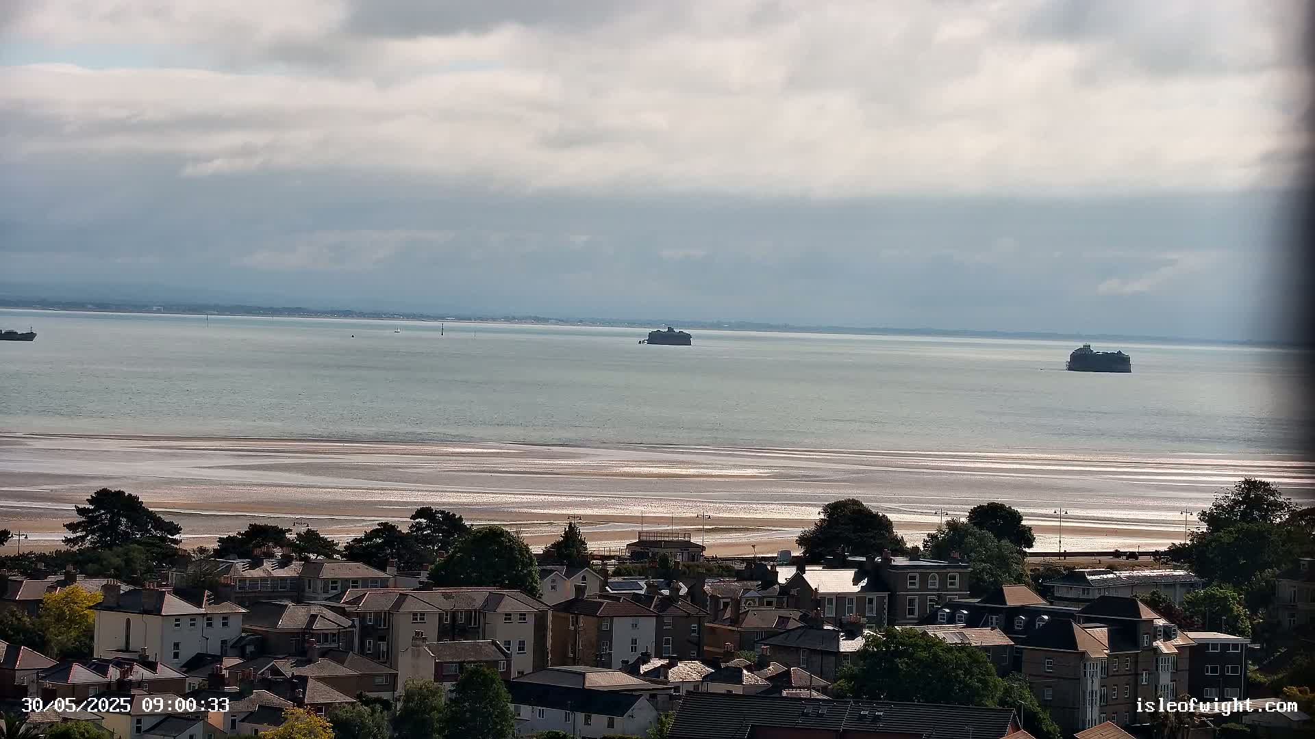 A coastal town overlooks a wide expanse of calm, light-grey water and a sandy beach under a mostly cloudy sky; two distant structures are visible in the water.