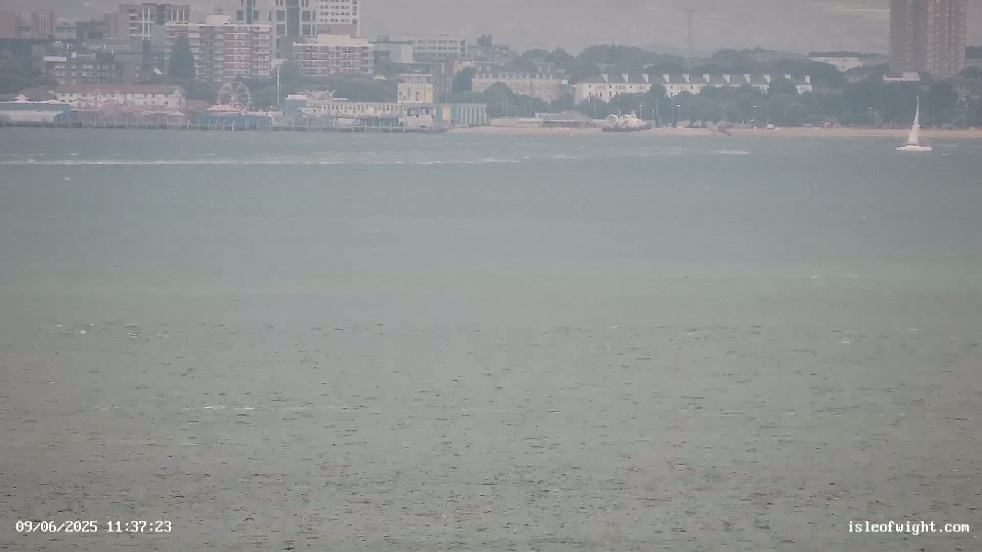 A hazy view of a body of water shows a distant shoreline with buildings, a Ferris wheel, and a sailboat under overcast skies.