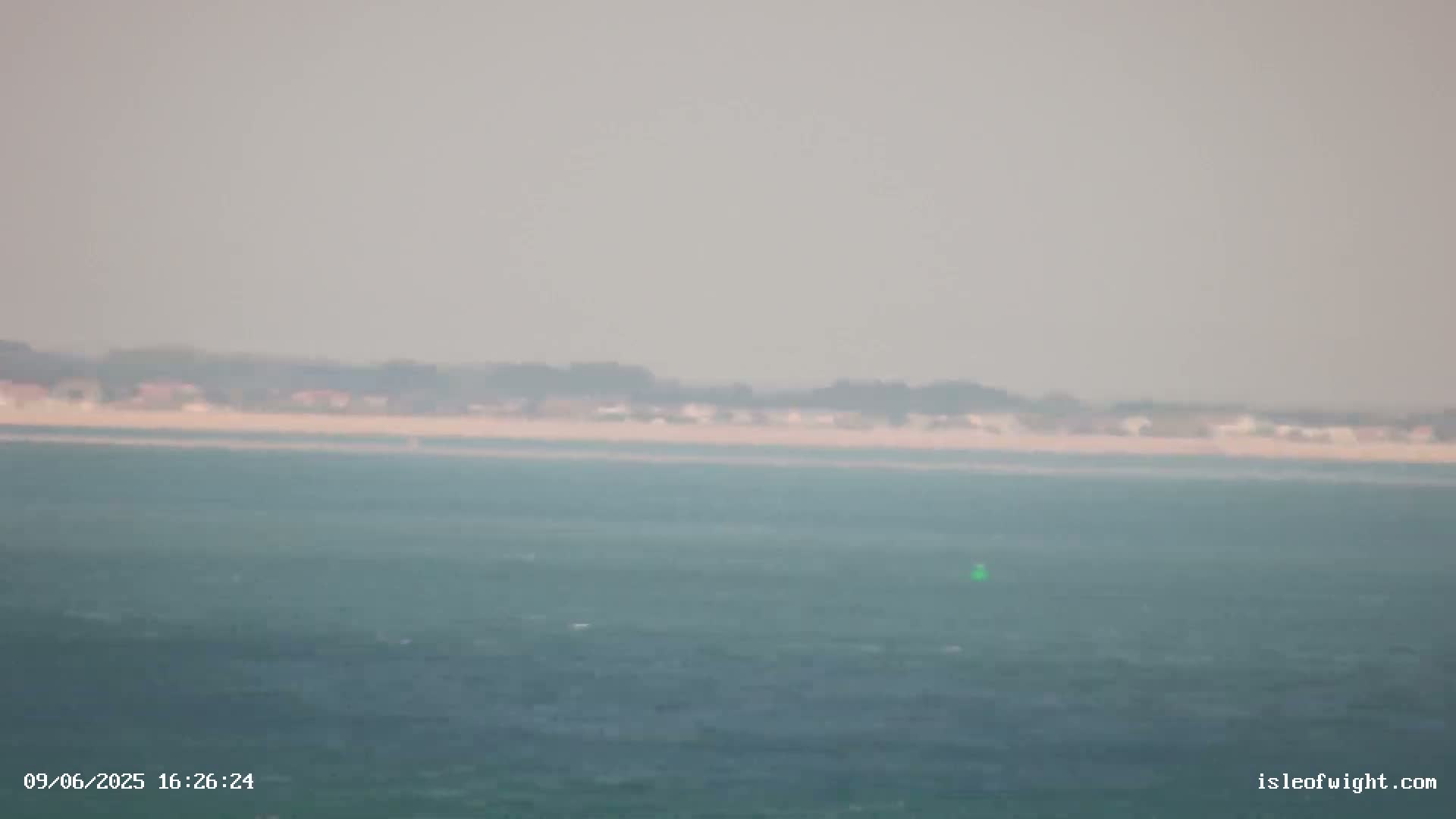 A hazy view shows a calm body of water with a distant shoreline featuring low-lying vegetation and buildings.