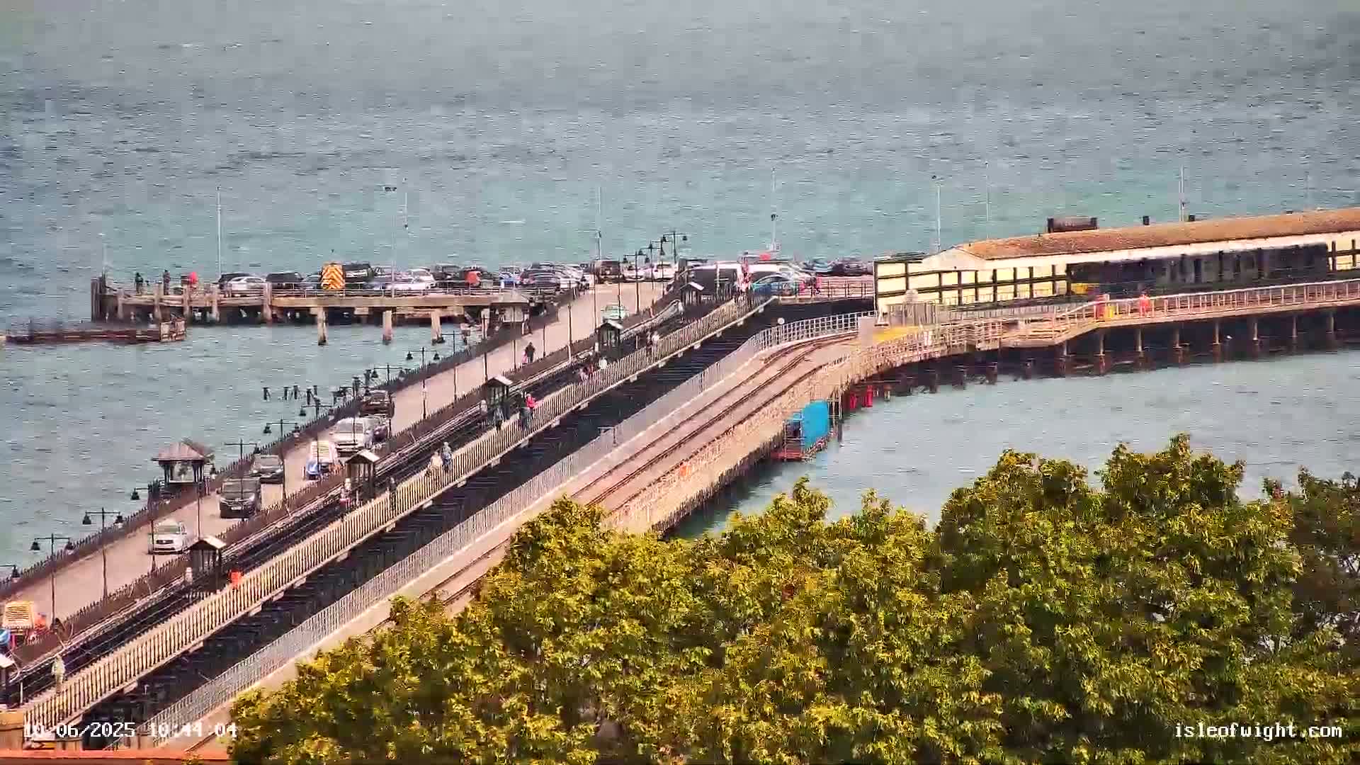 A long pier extends over a calm body of water, with vehicles and pedestrians visible on its surface.