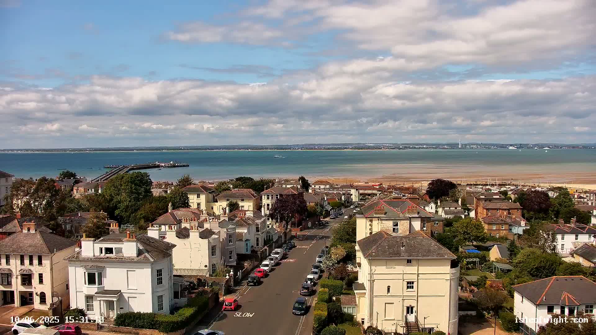 An elevated view of a seaside town on a partly cloudy day, showing residential streets and houses, a pier extending into the calm water of a bay, and a sandy beach in the distance.