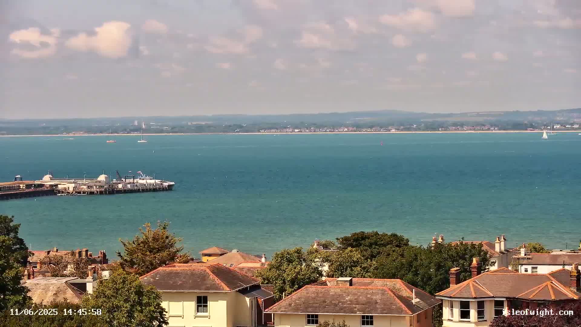 A waterfront view shows a body of teal water with a pier and several boats, under a partly cloudy sky, overlooking a collection of houses with brown roofs.