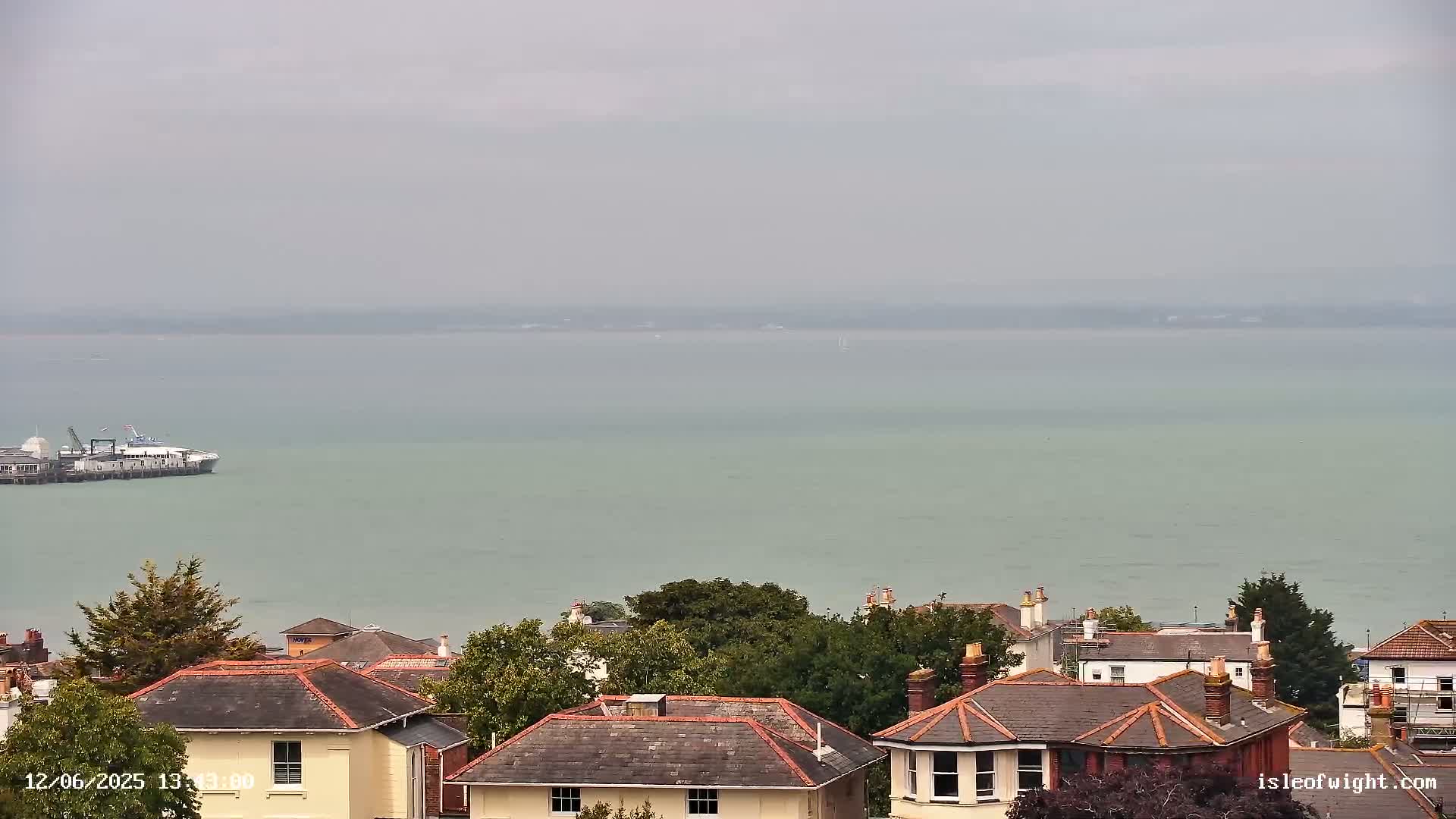 A ferry is docked near a shoreline of houses with gray, overcast skies above a calm, light-green body of water.