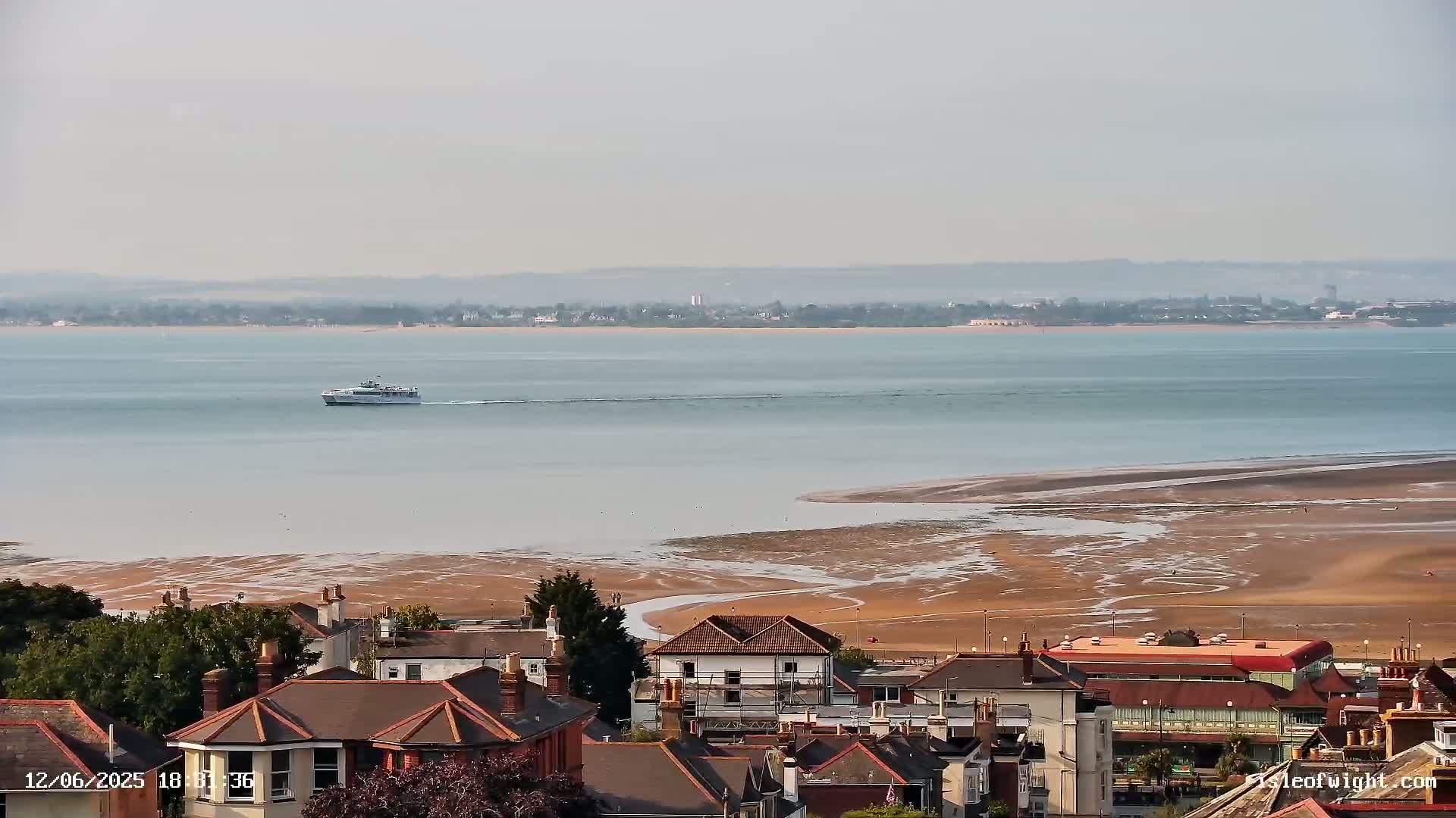 A ferry travels across a calm body of water, with a wide expanse of exposed sand visible at low tide in the foreground, and a town of red-roofed houses situated on a hill overlooking the water.