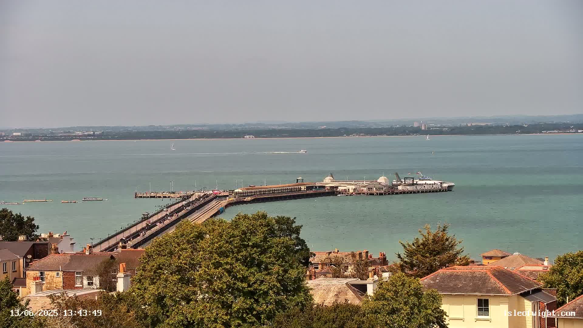 A long pier extends into a calm, light-green body of water on a hazy day, with buildings and trees visible in the foreground.