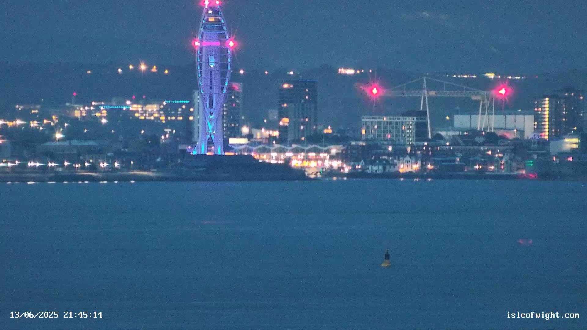 A brightly lit, slender tower stands on a waterfront at night, overlooking a city with many lights reflecting on dark water.
