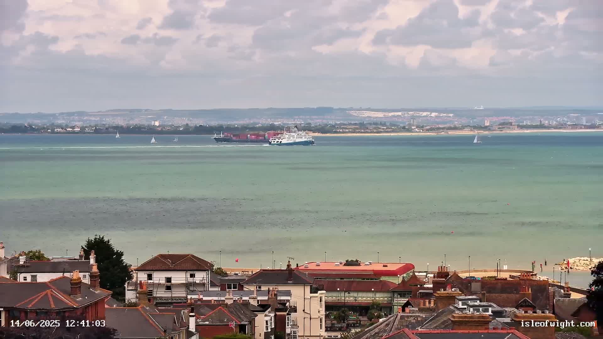 A coastal town overlooks a calm, light-green body of water where a large ferry and several sailboats are visible under a cloudy sky.