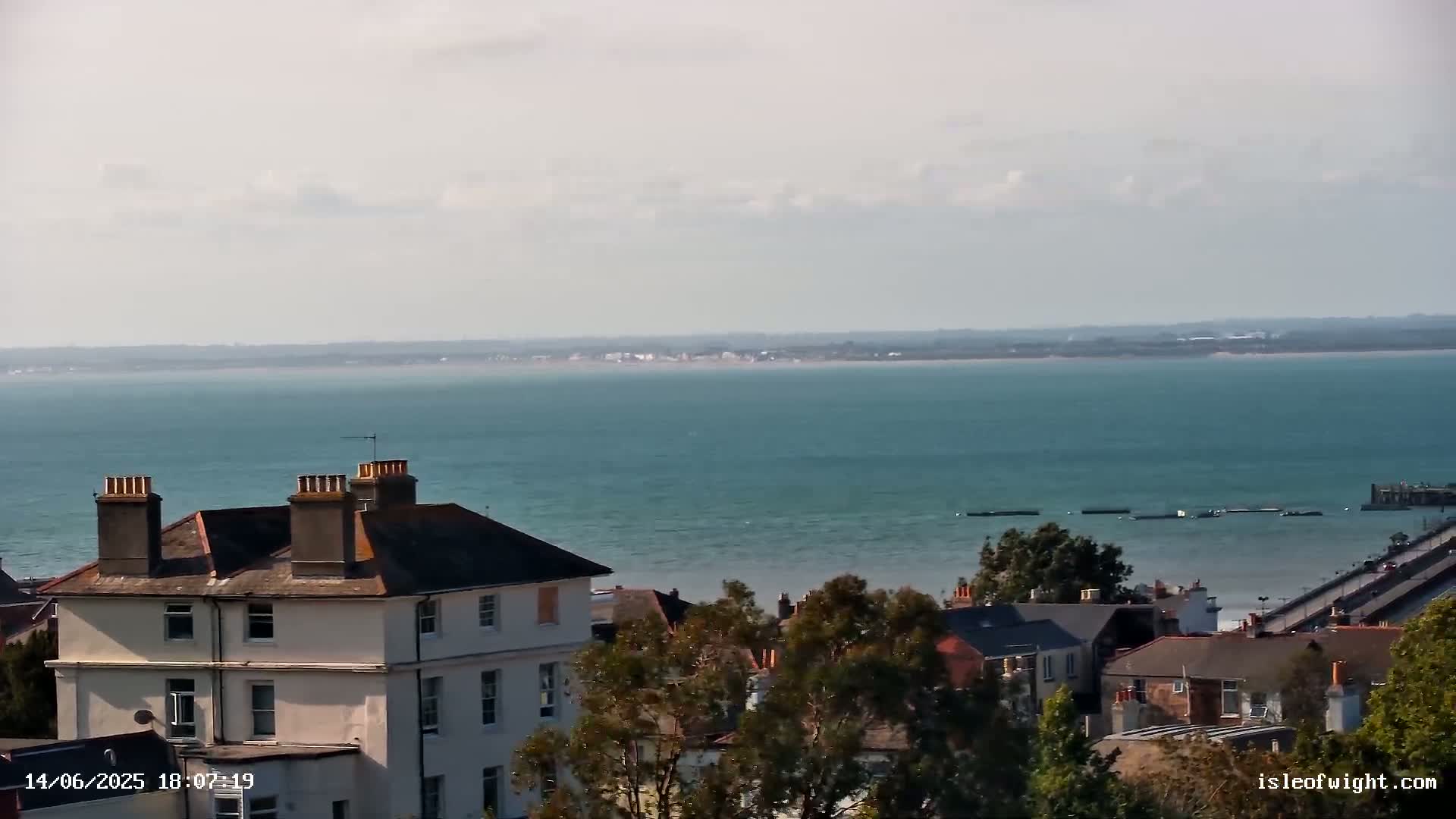 A seaside town's rooftops and trees overlook a calm, teal ocean under a mostly cloudy sky.