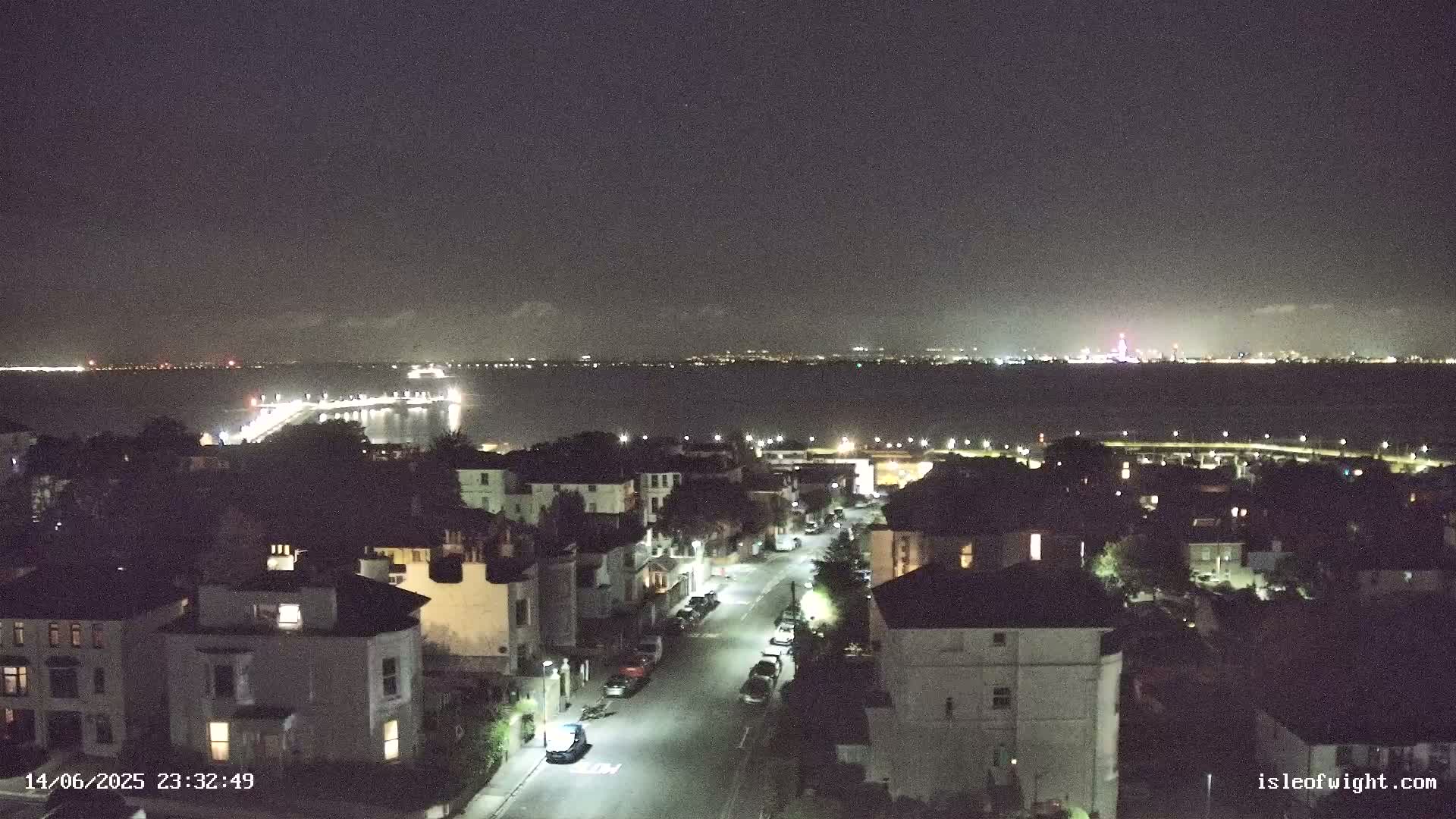 A seaside town's rooftops and trees overlook a calm, teal ocean under a mostly cloudy sky.