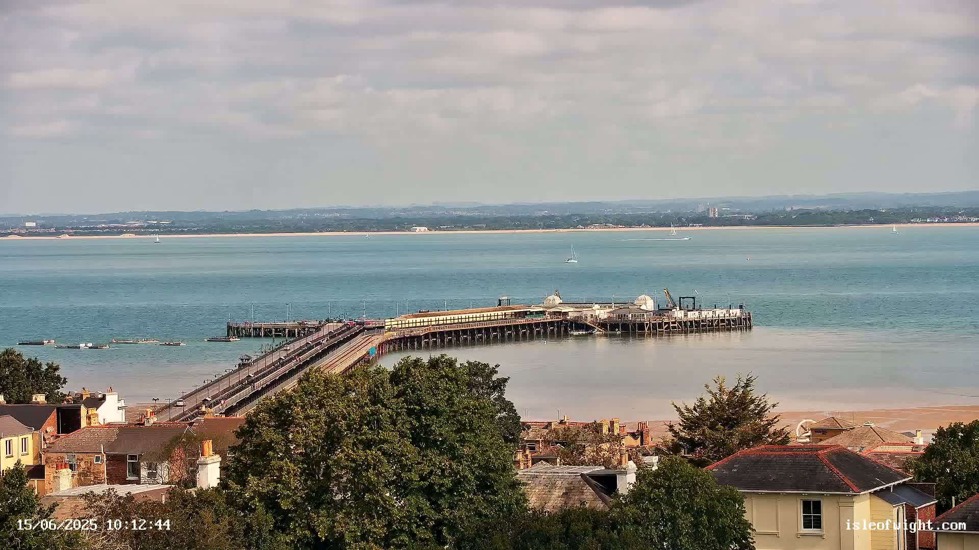 A long pier extends into a calm body of water under a partly cloudy sky, with a town visible in the foreground.