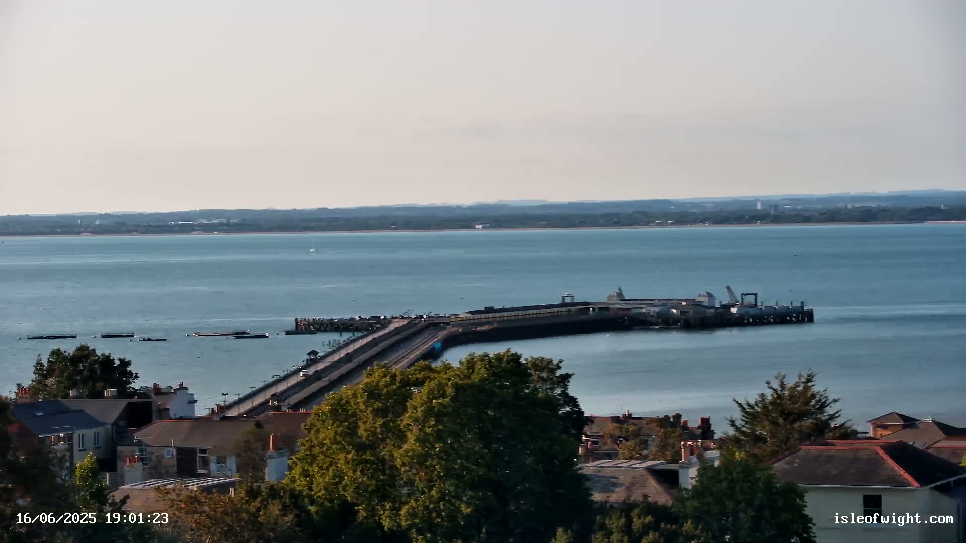 A long pier extends into a calm body of water under a clear sky, with buildings and trees visible in the foreground.