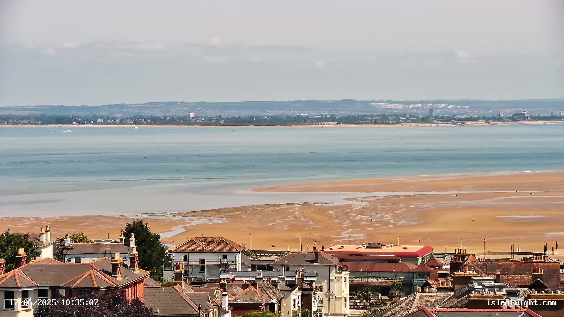 A coastal town overlooks a wide expanse of calm, light-blue water and a sandy beach at low tide under a mostly cloudy sky.