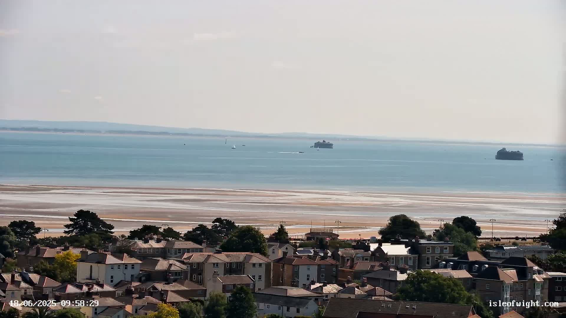 A coastal town overlooks a calm, light-blue sea with exposed sandbars at low tide, and two distant sea forts are visible on the horizon under a hazy sky.