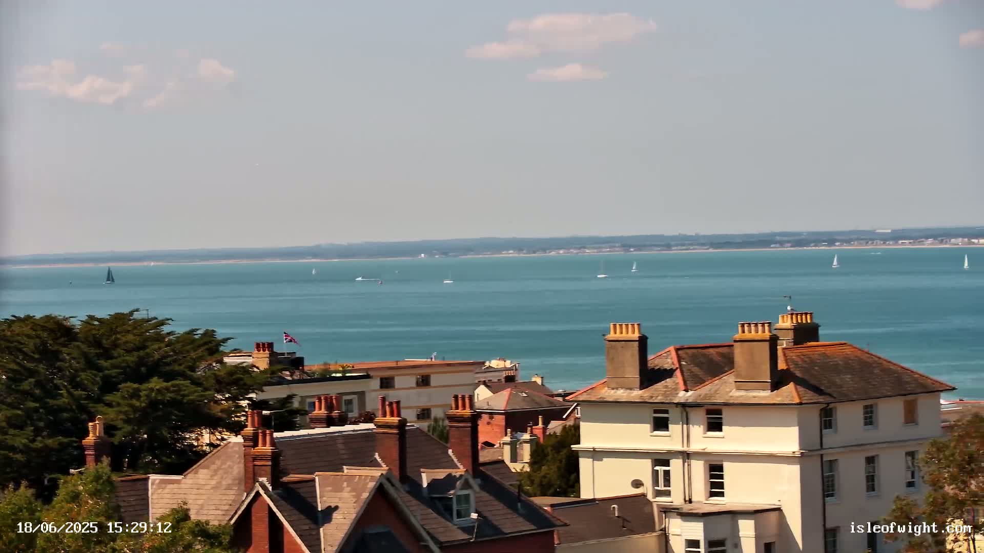 A view of several houses with brown roofs and chimneys overlooking a calm, blue body of water with sailboats on a sunny day.