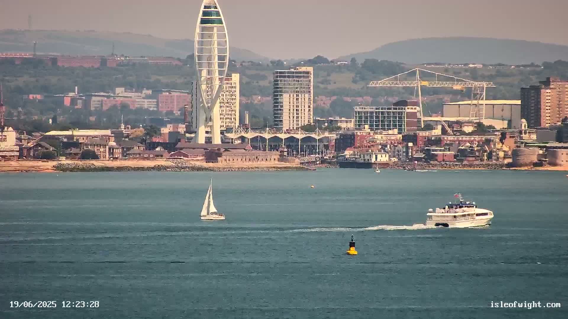 A sailboat and a ferry are on a calm body of water in front of a city skyline on a hazy day, with a tall, slender tower as a prominent feature.