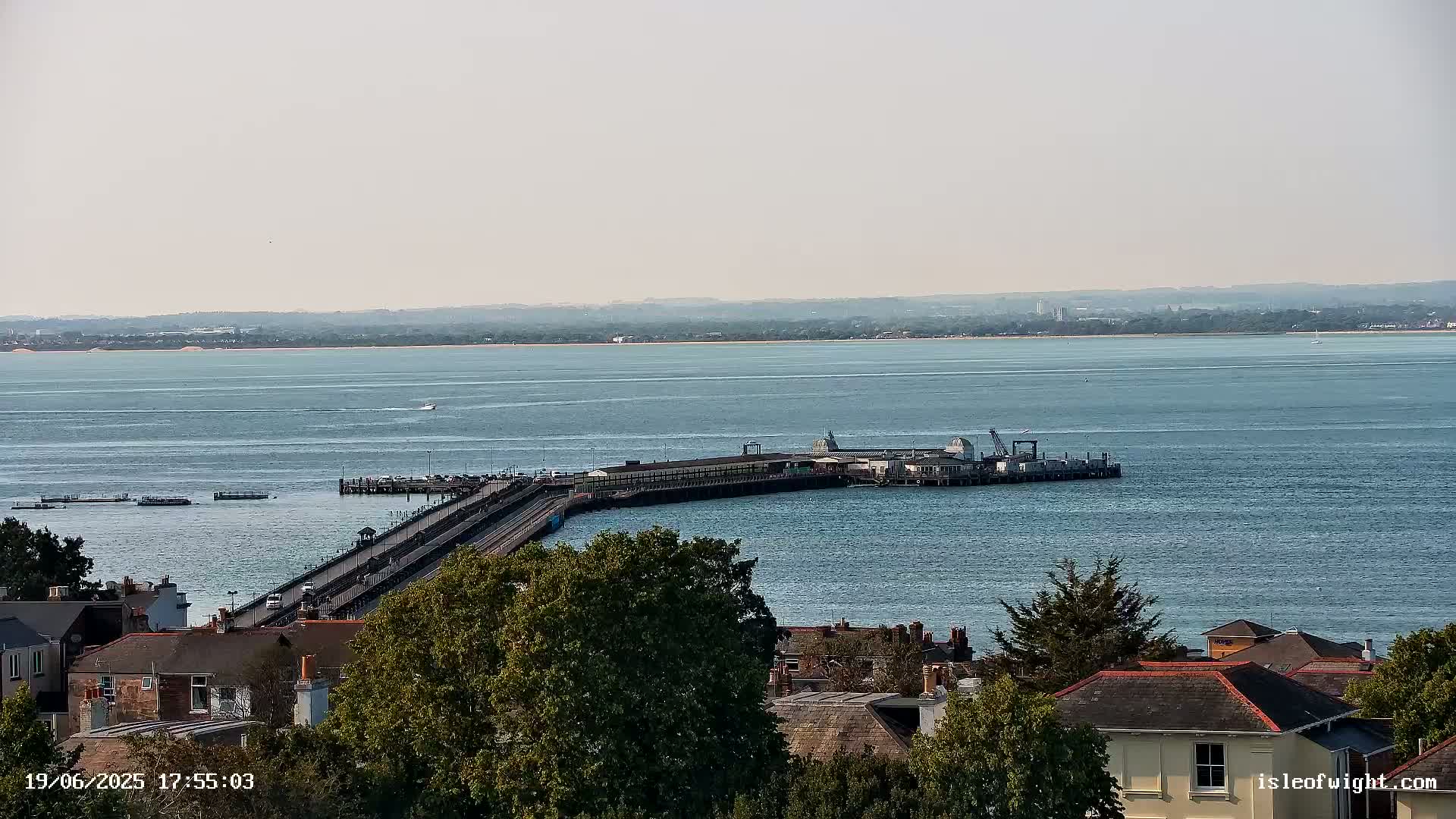 A long pier extends from a town into a calm, blue body of water, with a distant shoreline visible under a hazy sky.