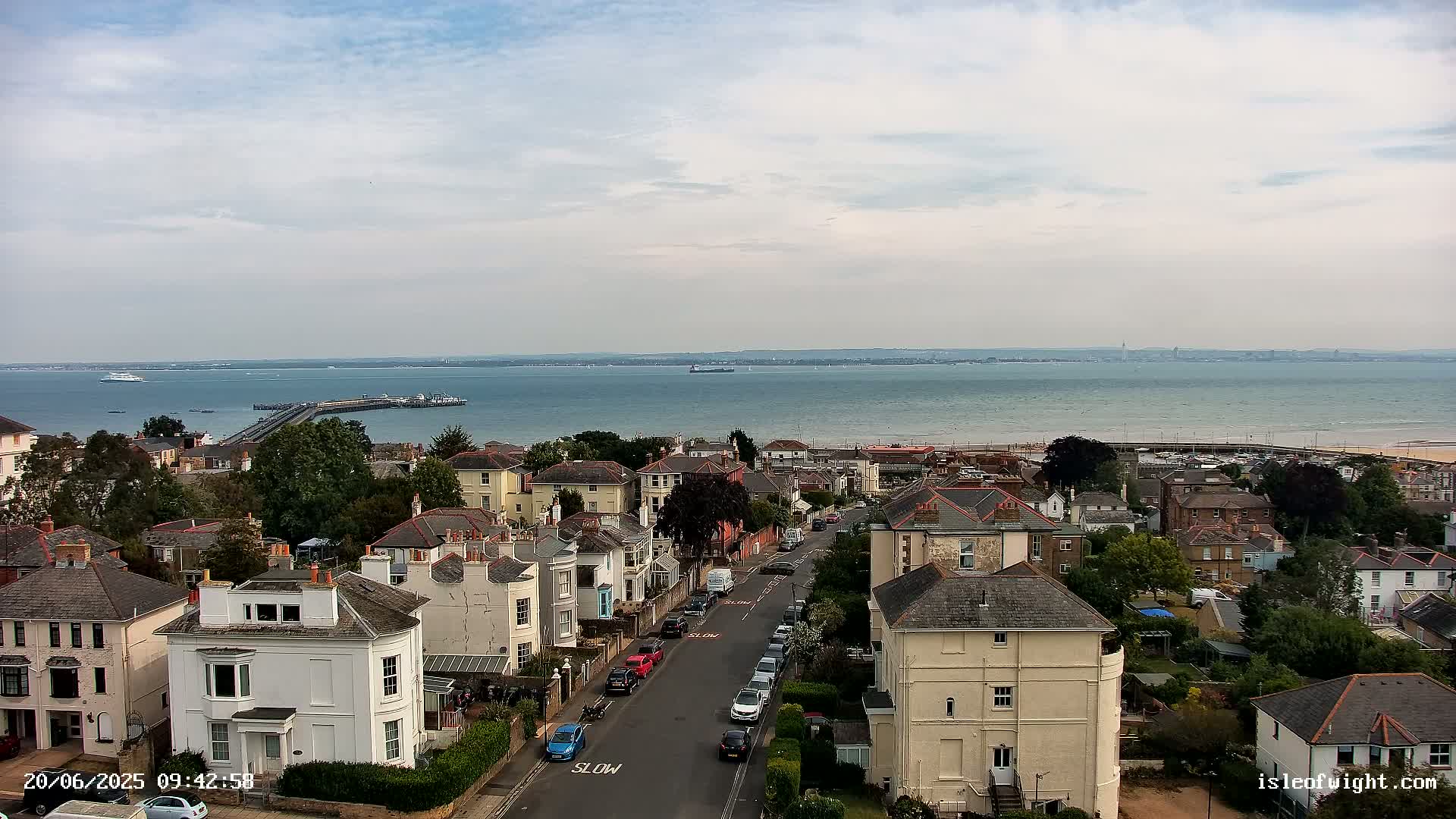 A coastal town with two-story houses and a long pier extending into a calm, light-blue sea under a partly cloudy sky.