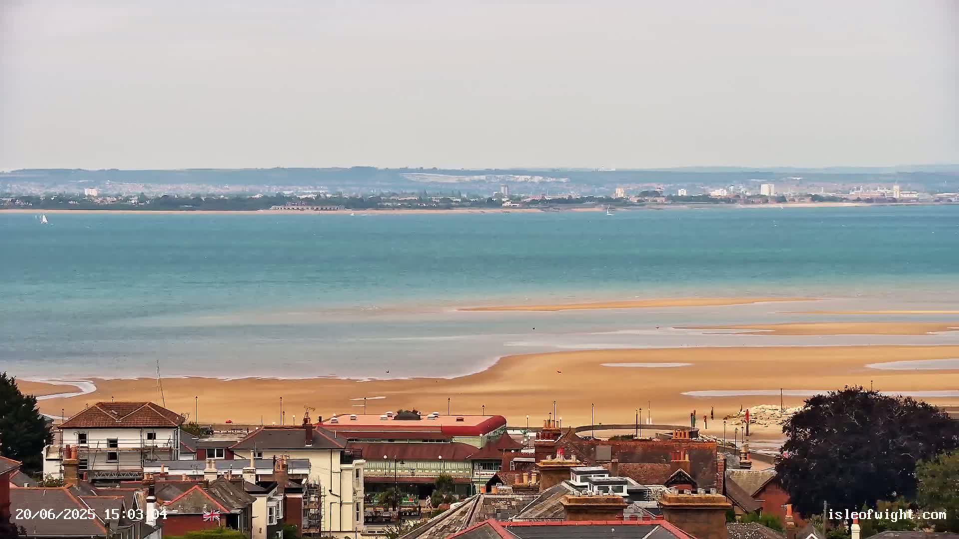 A coastal town overlooks a wide expanse of calm, teal water and a large, pale sandbar at low tide, with a distant city skyline visible on the horizon under an overcast sky.