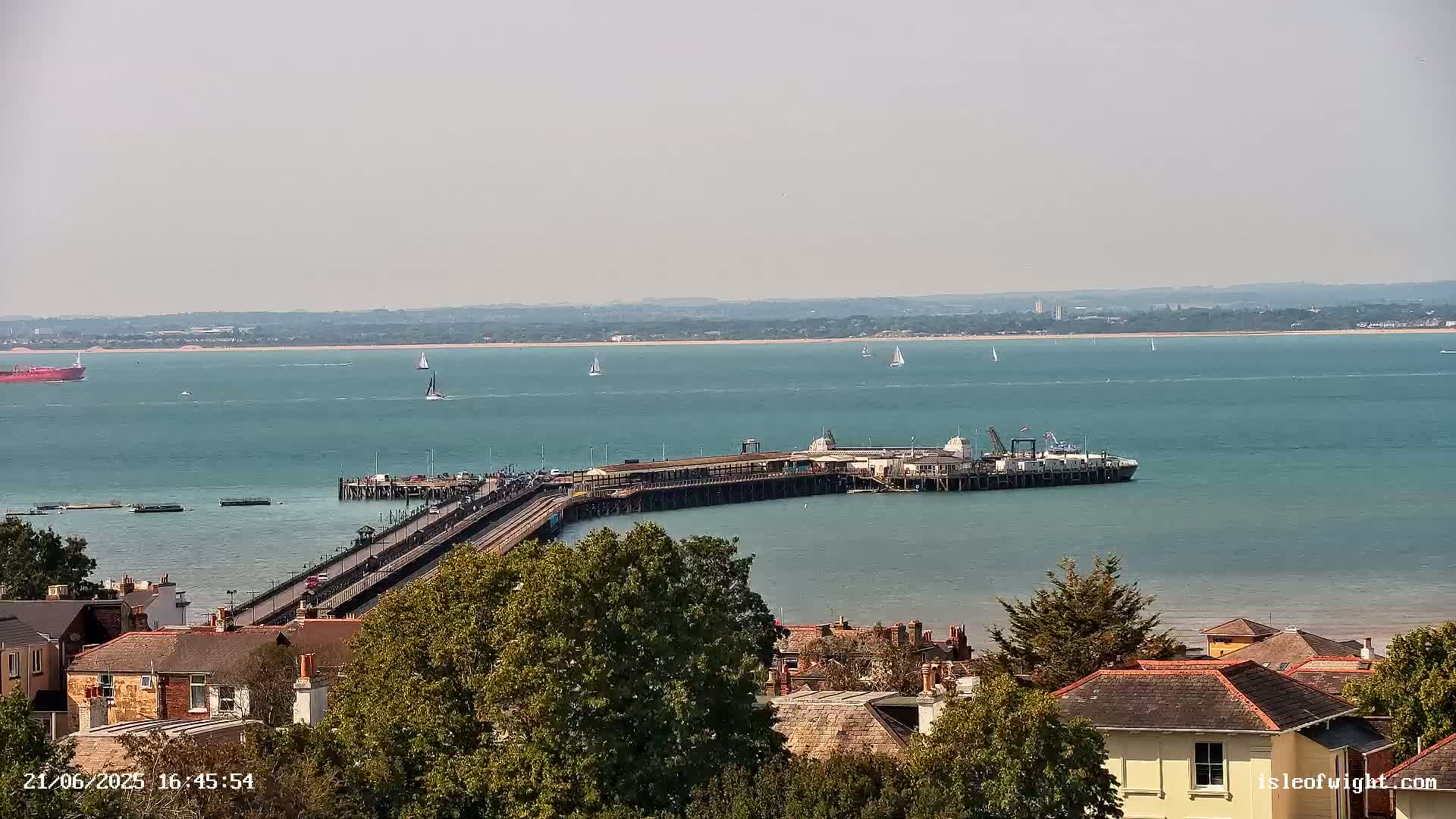 A long pier extends into a calm, teal ocean, with several sailboats and a large cargo ship visible in the distance, under a hazy sky.