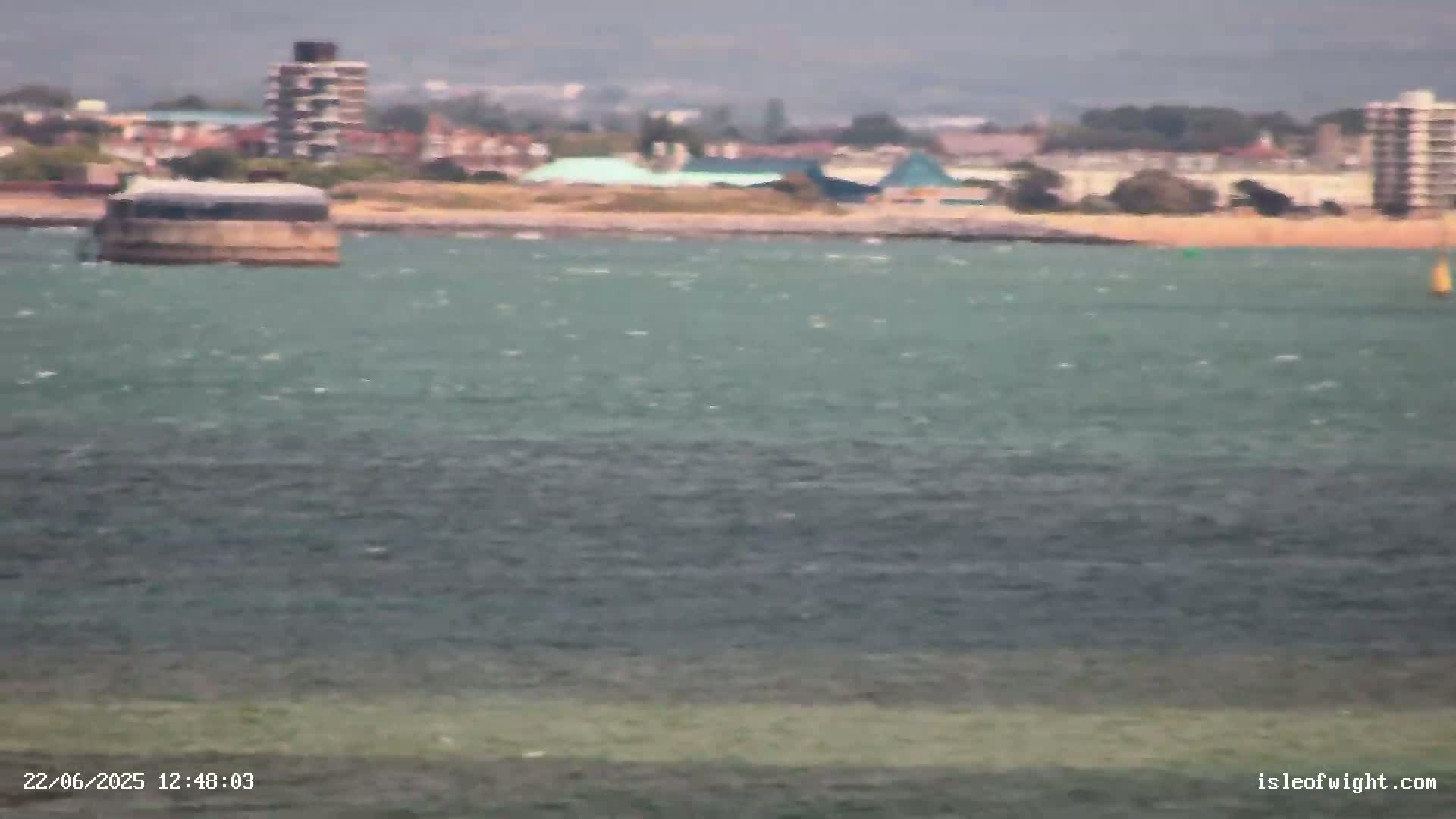 A low-lying structure sits in a choppy body of water in front of a distant shoreline with buildings under an overcast sky.