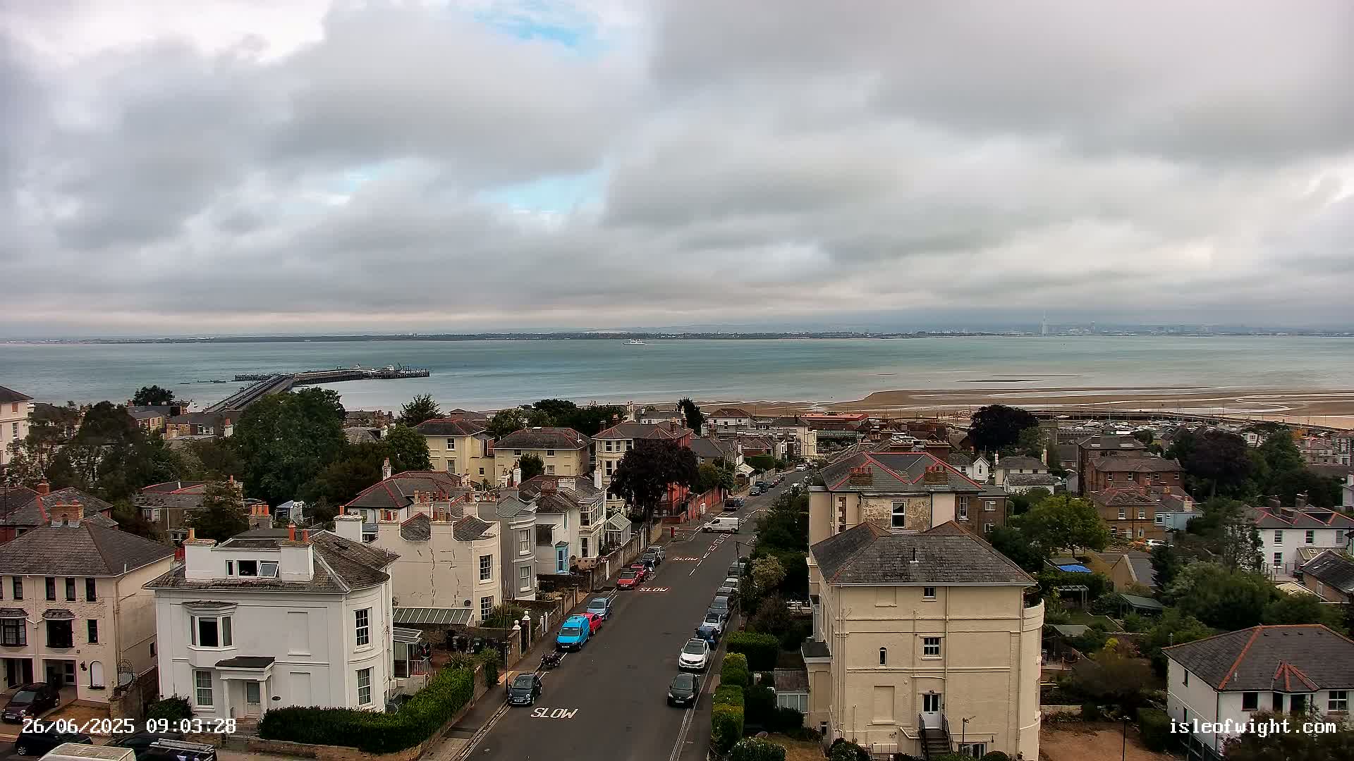 A coastal town with houses and a street, overlooks a calm body of water under a cloudy sky.