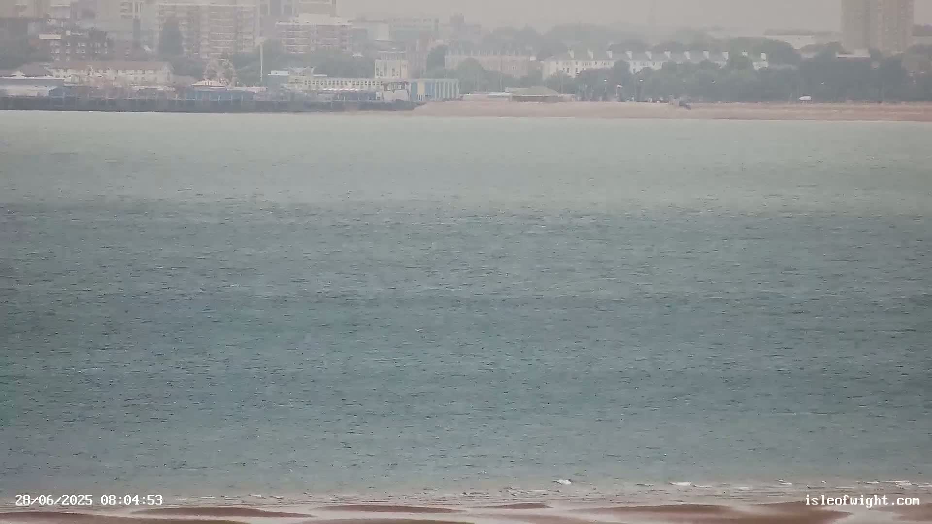 A hazy, overcast day shows a calm body of water with a distant shoreline featuring buildings and a beach.