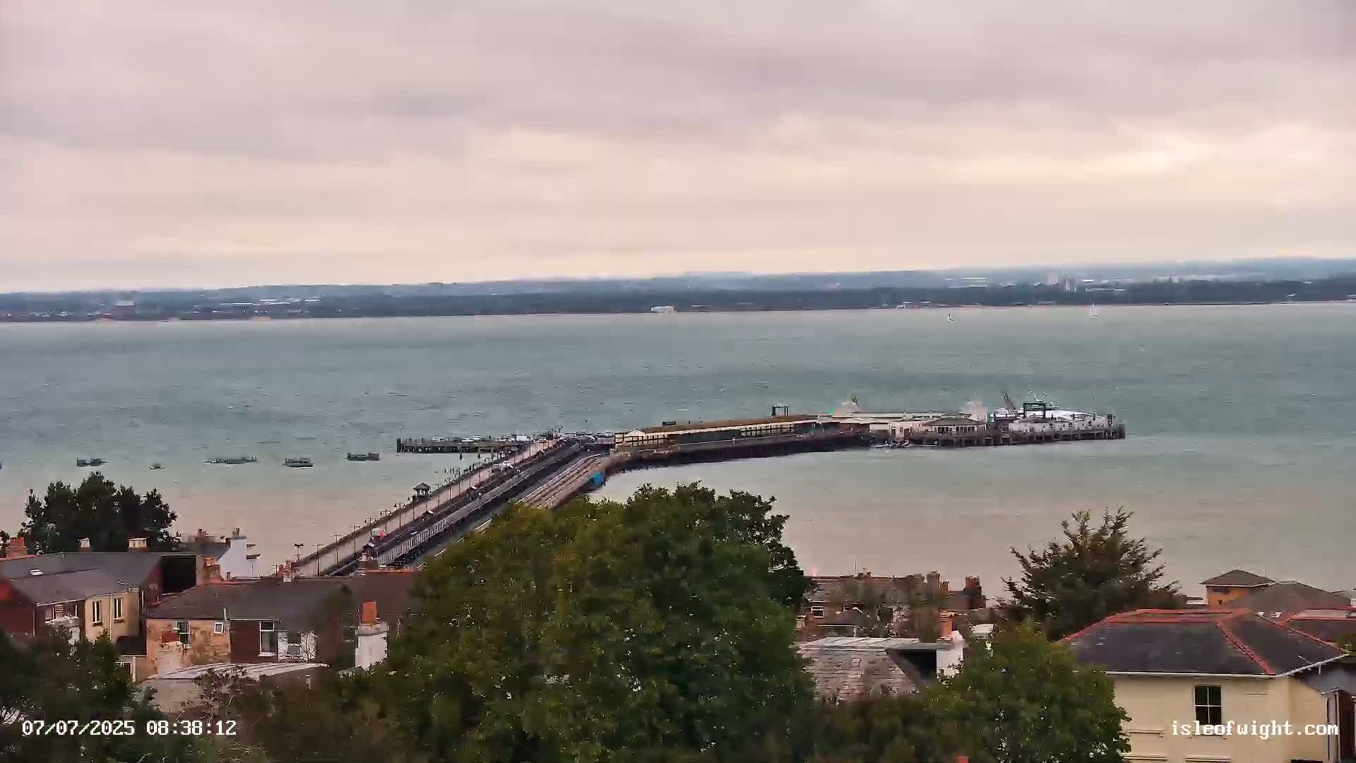 A long pier extends into a calm, gray body of water under an overcast sky, with residential buildings visible in the foreground.