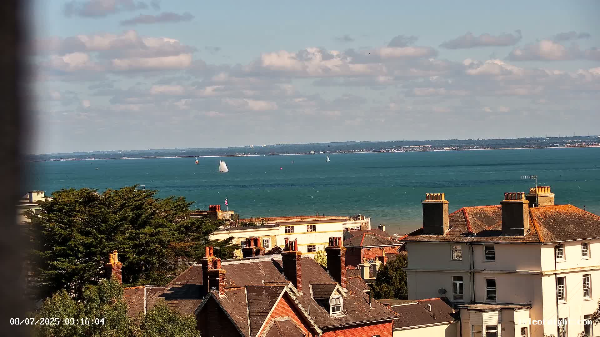 A partly cloudy day reveals a seaside town with several houses and sailboats visible on the calm blue water in the distance.