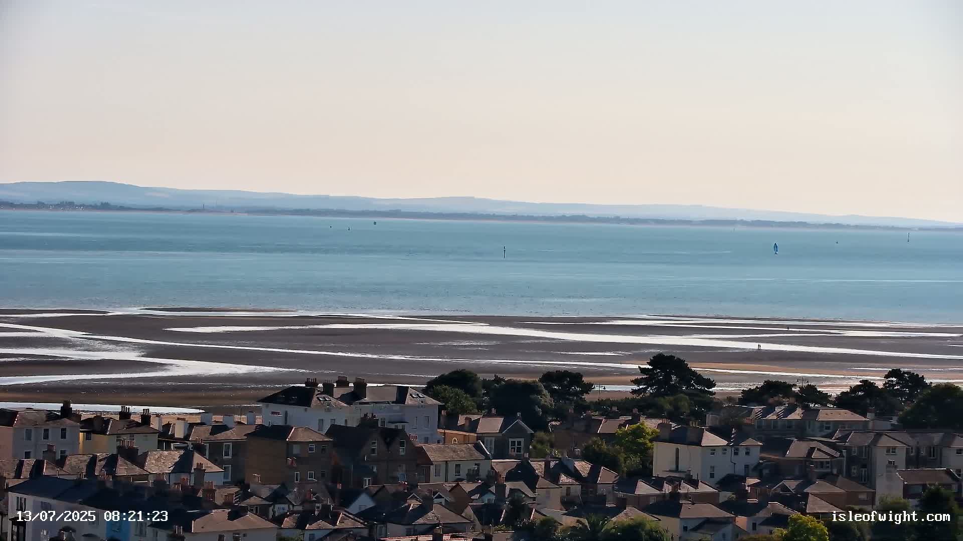 A coastal town overlooks a wide expanse of calm, blue water and a flat, dark sand beach at low tide under a clear sky.
