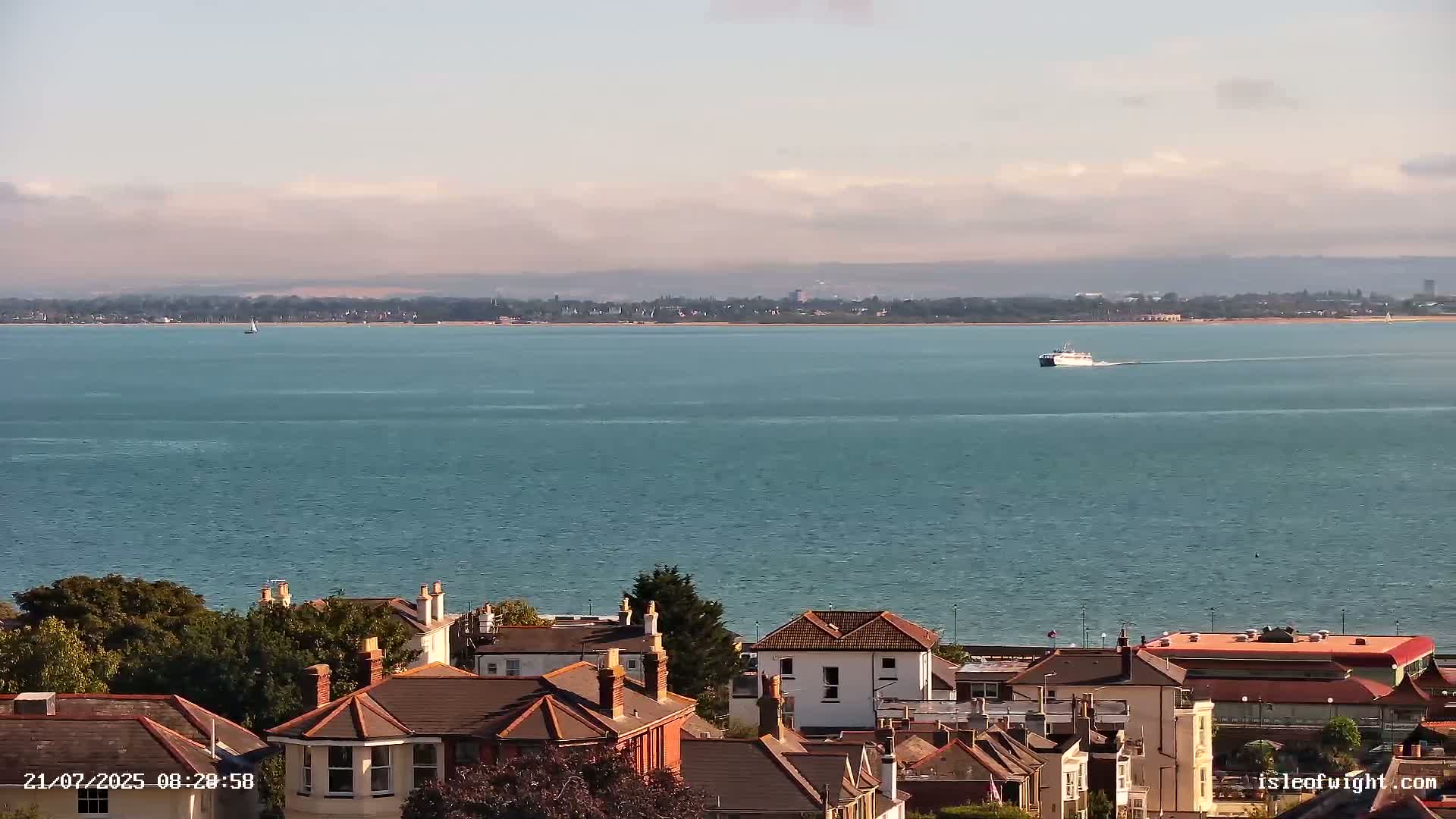 A calm, blue body of water stretches to a distant shoreline,  a ferry and sailboat visible on the water, with a collection of houses and trees in the foreground under a mostly clear sky.