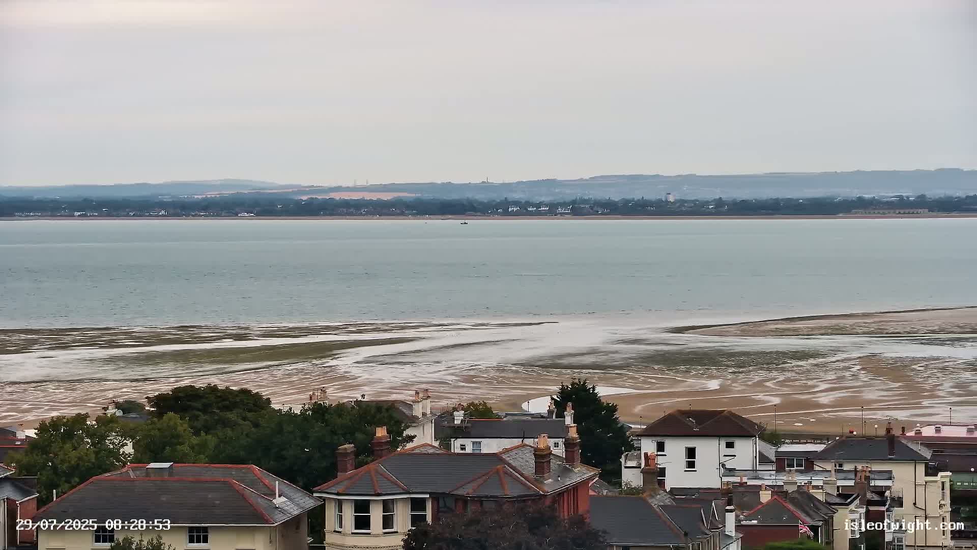 A coastal town overlooks a wide expanse of calm, grey water receding to reveal a sandy shore patterned with tidal channels under an overcast sky.
