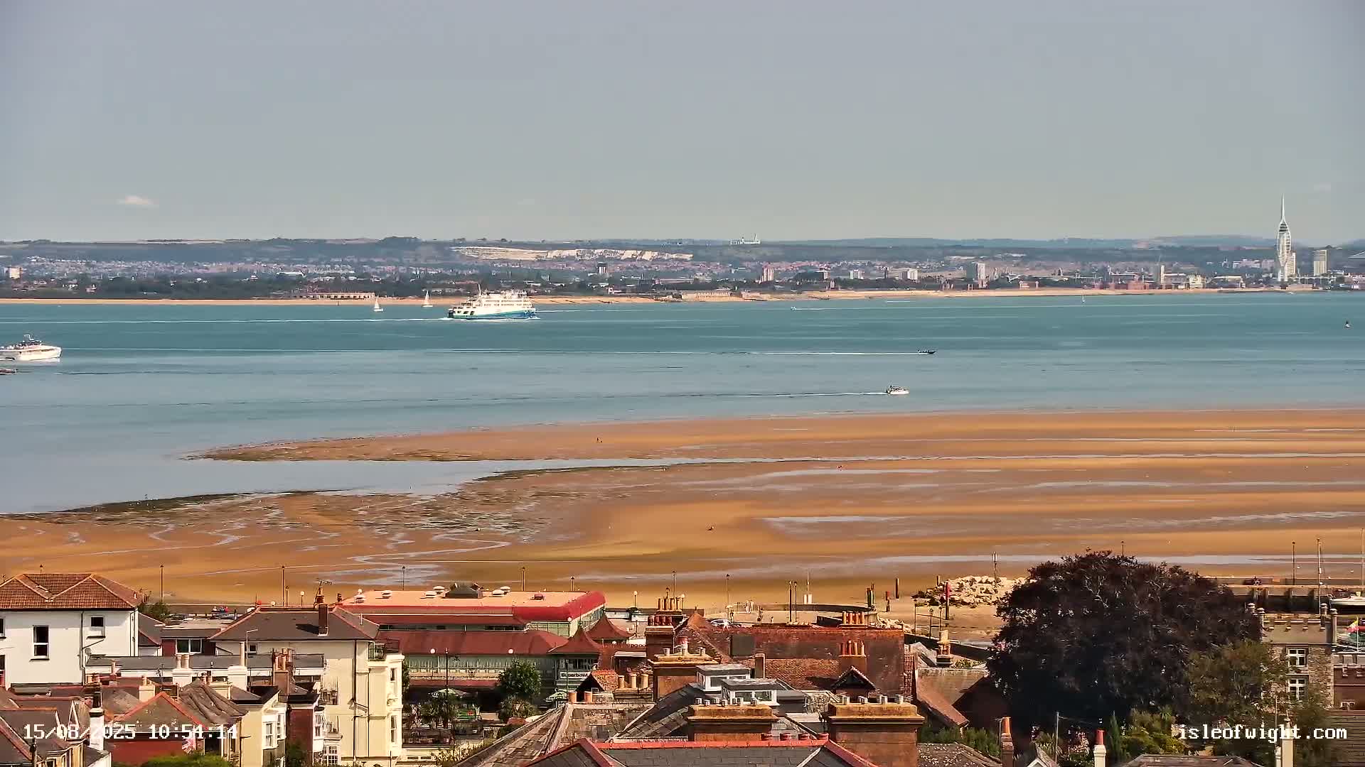 A coastal town overlooks a wide expanse of calm, grey water receding to reveal a sandy shore patterned with tidal channels under an overcast sky.