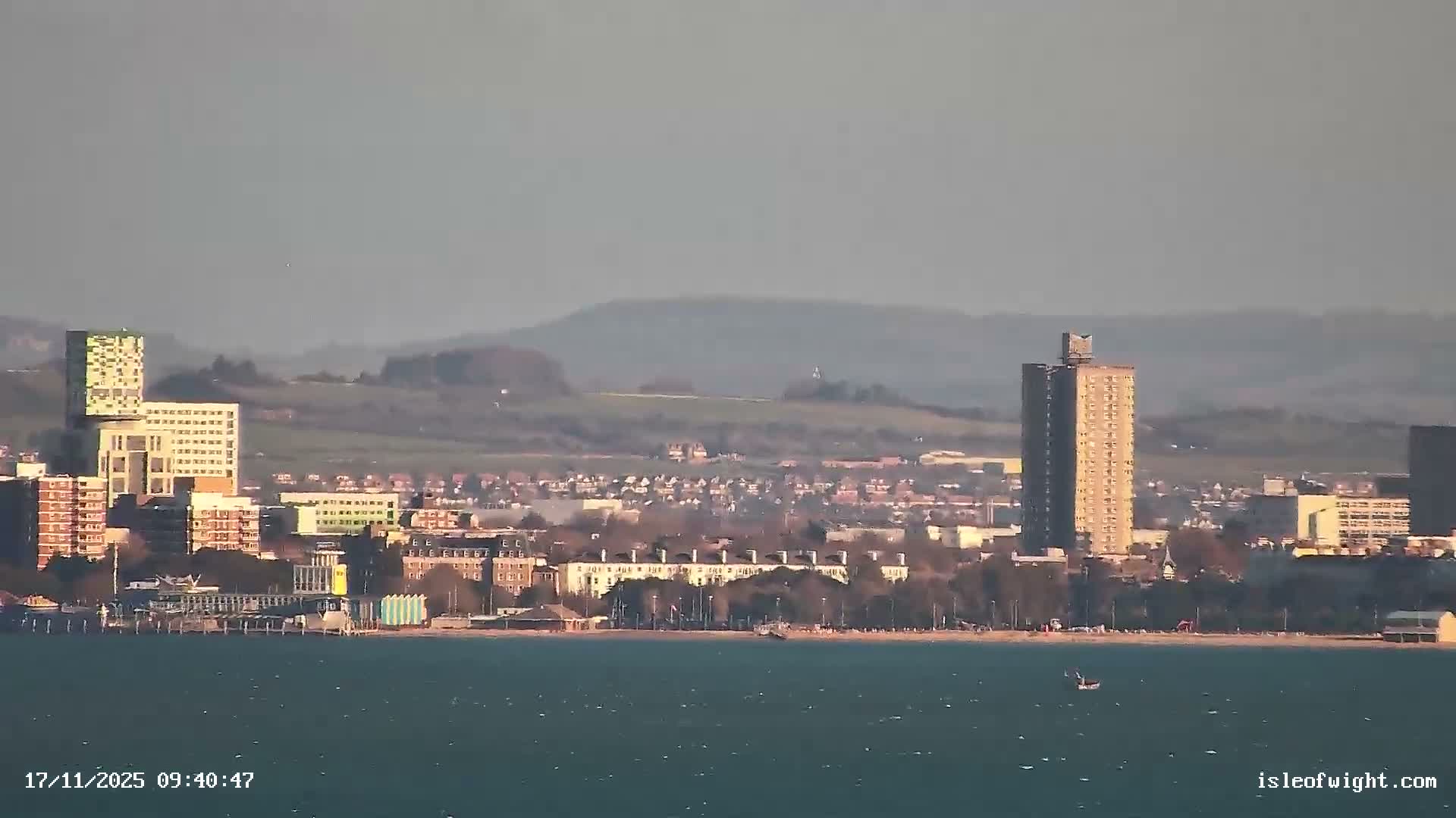 A coastal town sprawls along a choppy blue-green bay under clear, sunny skies, with diverse buildings, a beach, and distant rolling hills visible, and a small boat in the water.