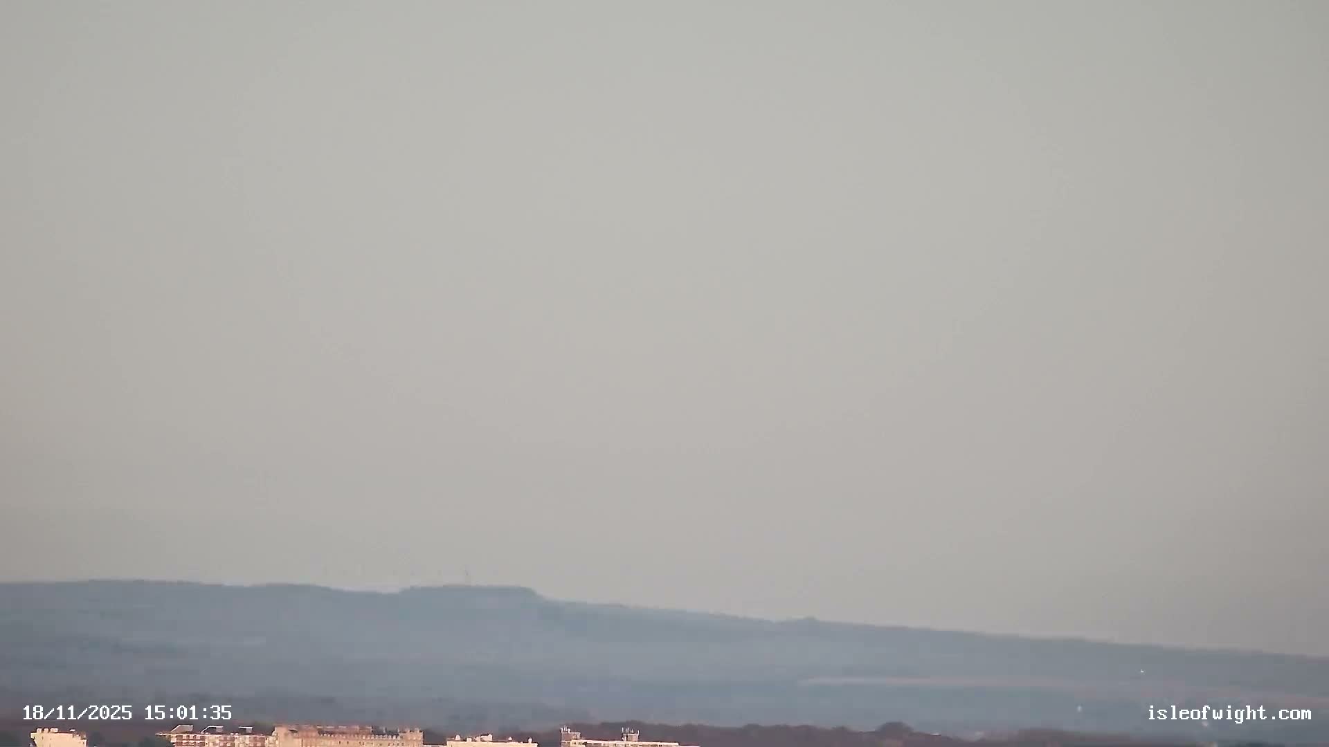 A coastal town sprawls along a choppy blue-green bay under clear, sunny skies, with diverse buildings, a beach, and distant rolling hills visible, and a small boat in the water.
