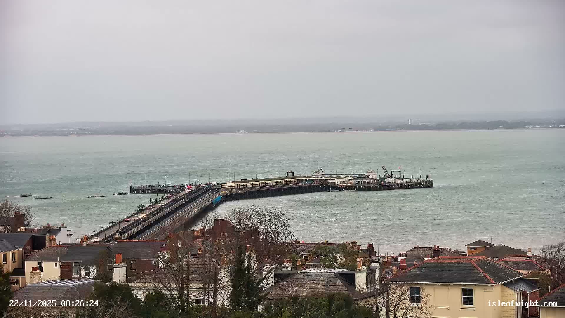 The image captures an overcast morning view of a long pier extending into choppy grey-green water, with residential buildings lining the shore in the foreground and a hazy landmass on the distant horizon.