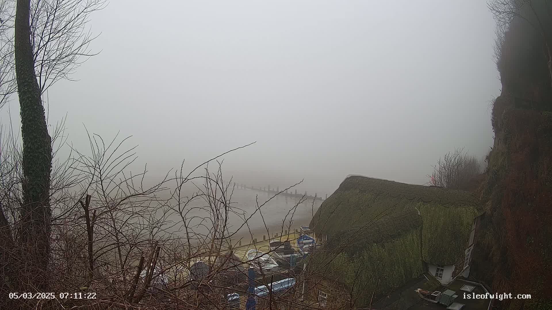 A foggy seaside scene is visible from a high vantage point, showing several small boats docked near a thatched-roof building situated on a cliffside.