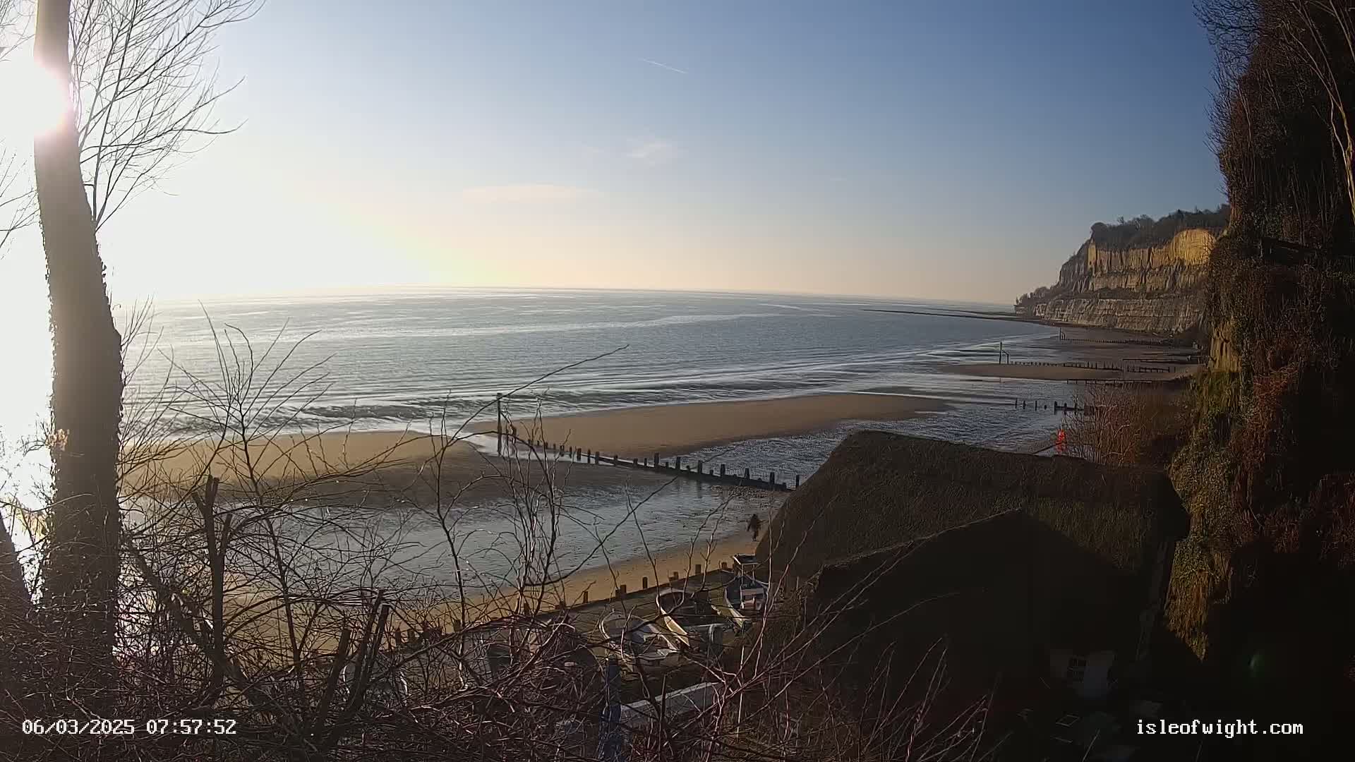 A sandy beach stretches below a high, light-brown cliff, with calm ocean waters under a mostly clear, sunny sky.
