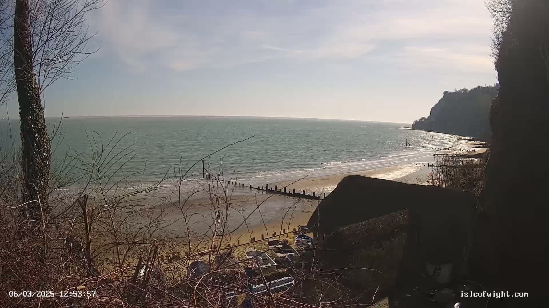 A sandy beach with calm ocean waves under a mostly sunny sky is viewed from a high vantage point, partially obscured by bare winter branches and a dark structure.