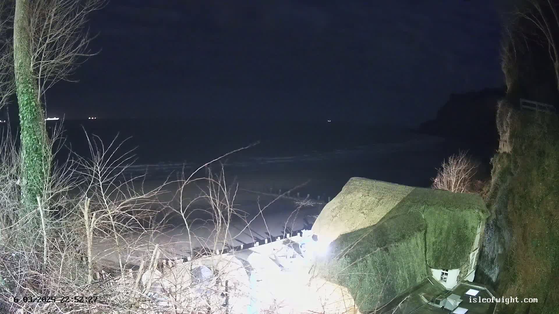 A dark, nighttime scene shows a thatched-roof building overlooking a beach and a calm, dark sea under a clear, black sky.